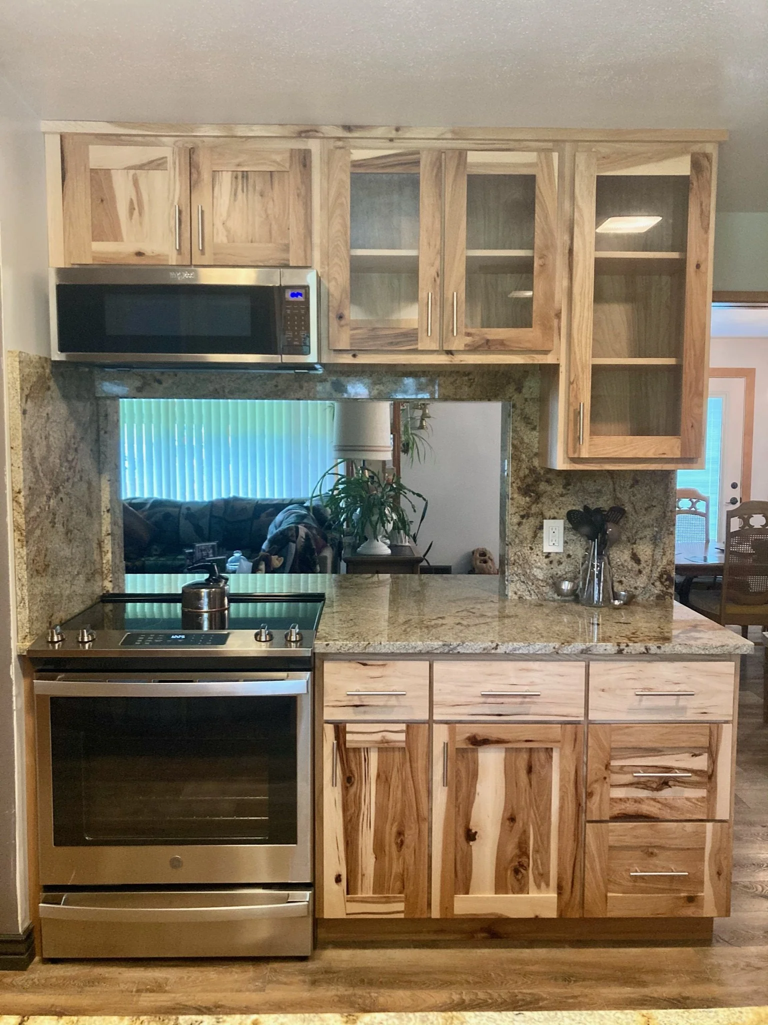 Kitchen view through an opening with granite countertops and wooden cabinets above and below, a stainless steel stove and microwave, and a glimpse into a living area with a couch, potted plant, and window with blinds.