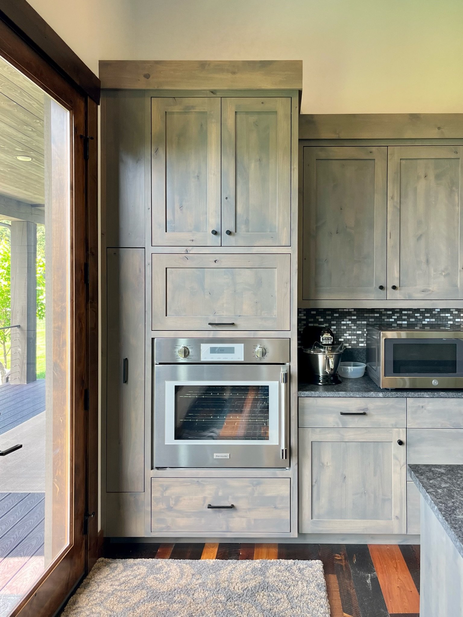 Kitchen with light wood cabinets, stainless steel oven, microwave, black tiled backsplash, and a door leading outside, with a beige rug on the floor.