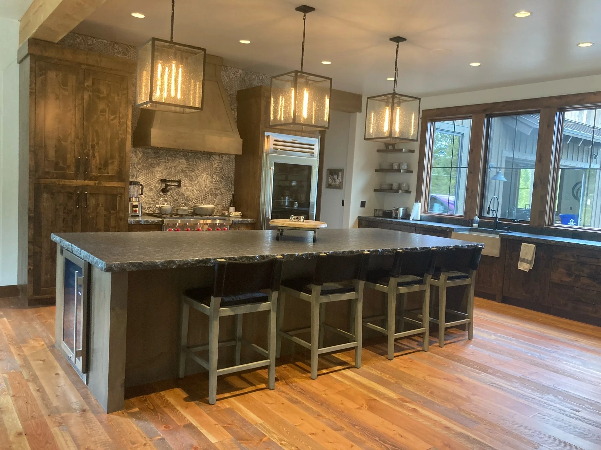 Modern kitchen with wooden cabinets, a large island with a dark countertop, and pendant lights hanging from the ceiling. The kitchen features large windows and open shelves with dishes.