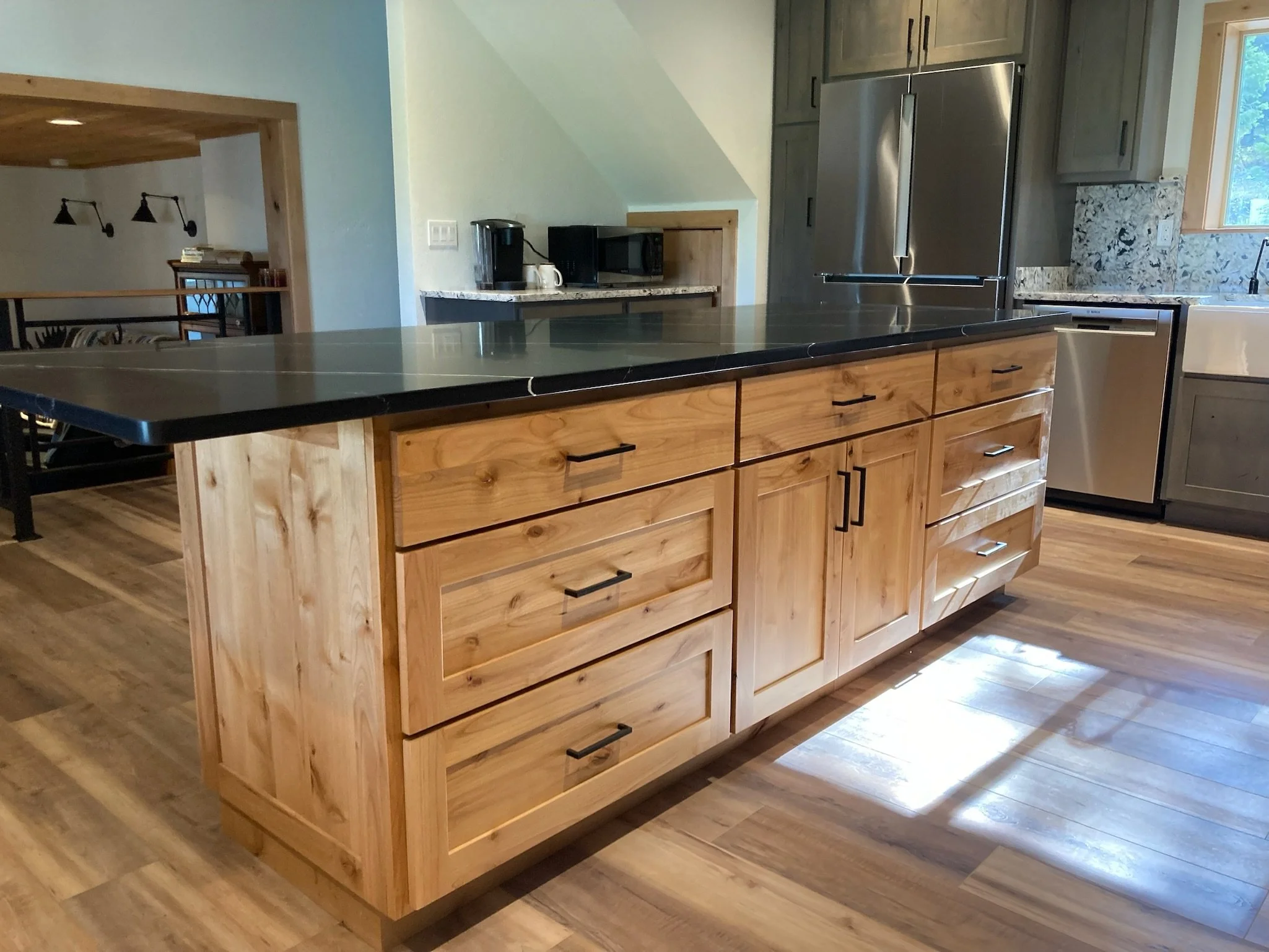 A kitchen island with a black countertop and wooden drawers, surrounded by cabinets, a refrigerator, and a window, with sunlight on the wooden floor.