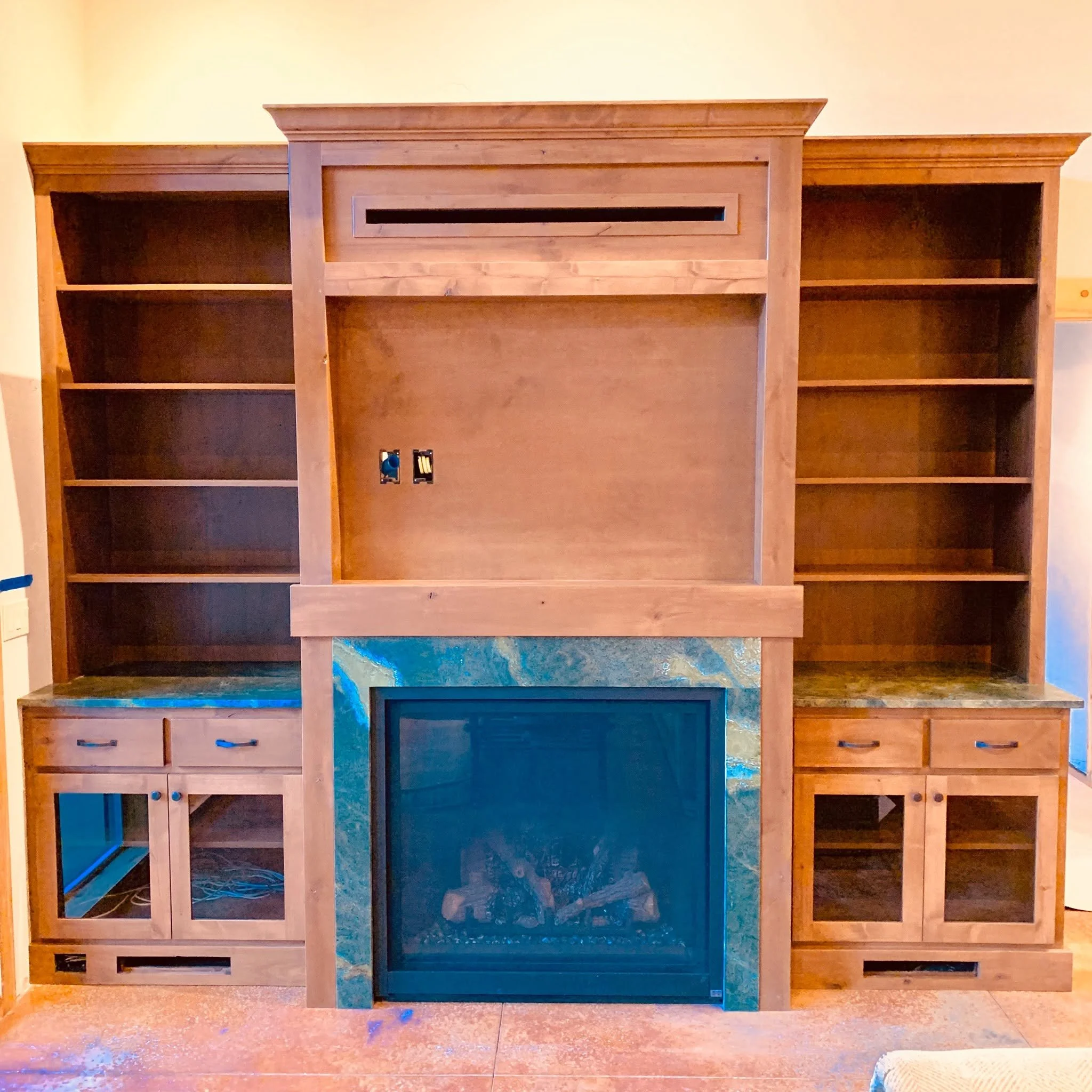Unfinished wooden cabinet or shelving unit with multiple open shelves and a centered space with electrical outlets, set against a beige wall.