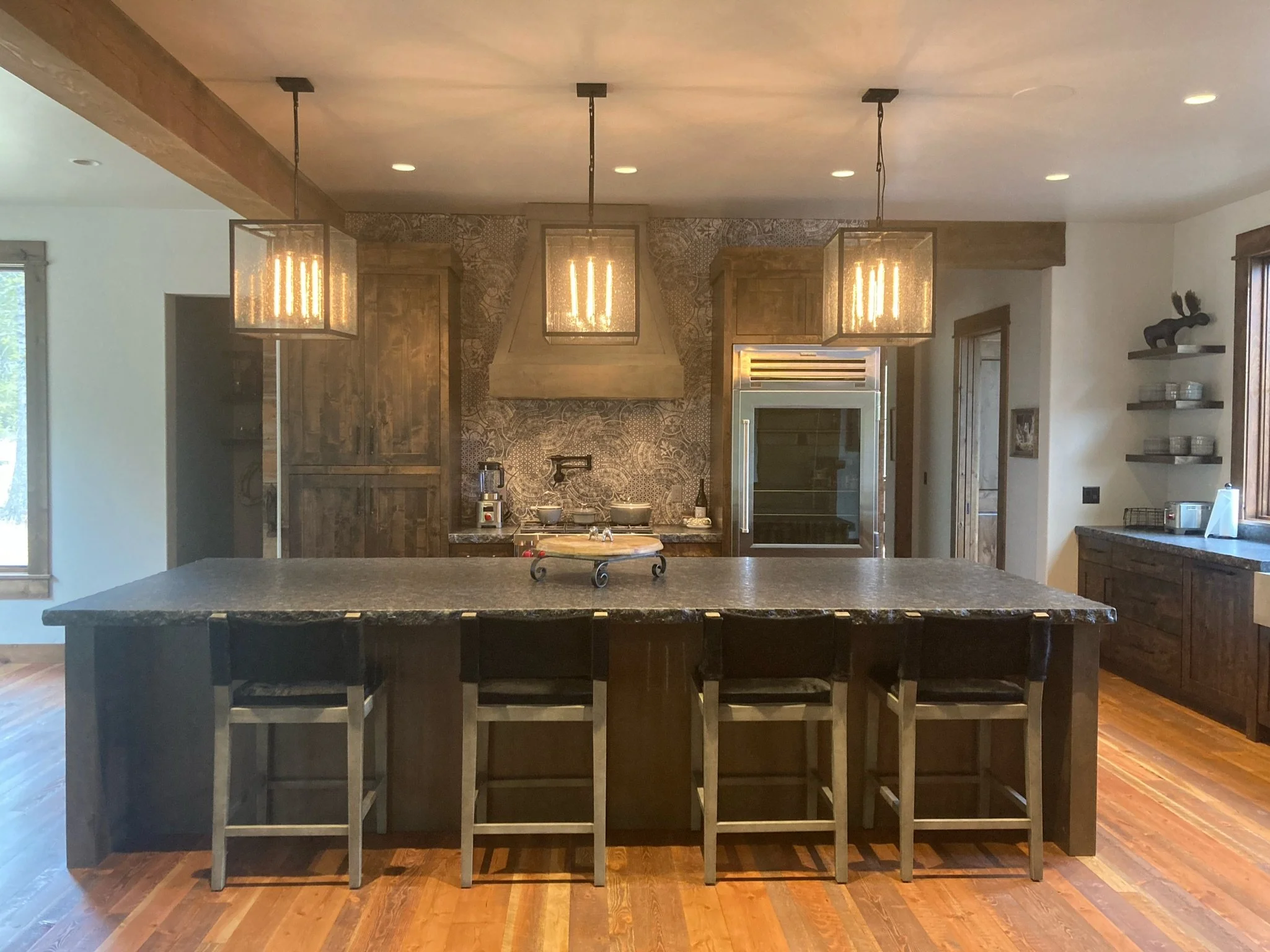 Modern kitchen with a large central island, hanging pendant lights, wooden cabinets, and stainless steel appliances, including a refrigerator.