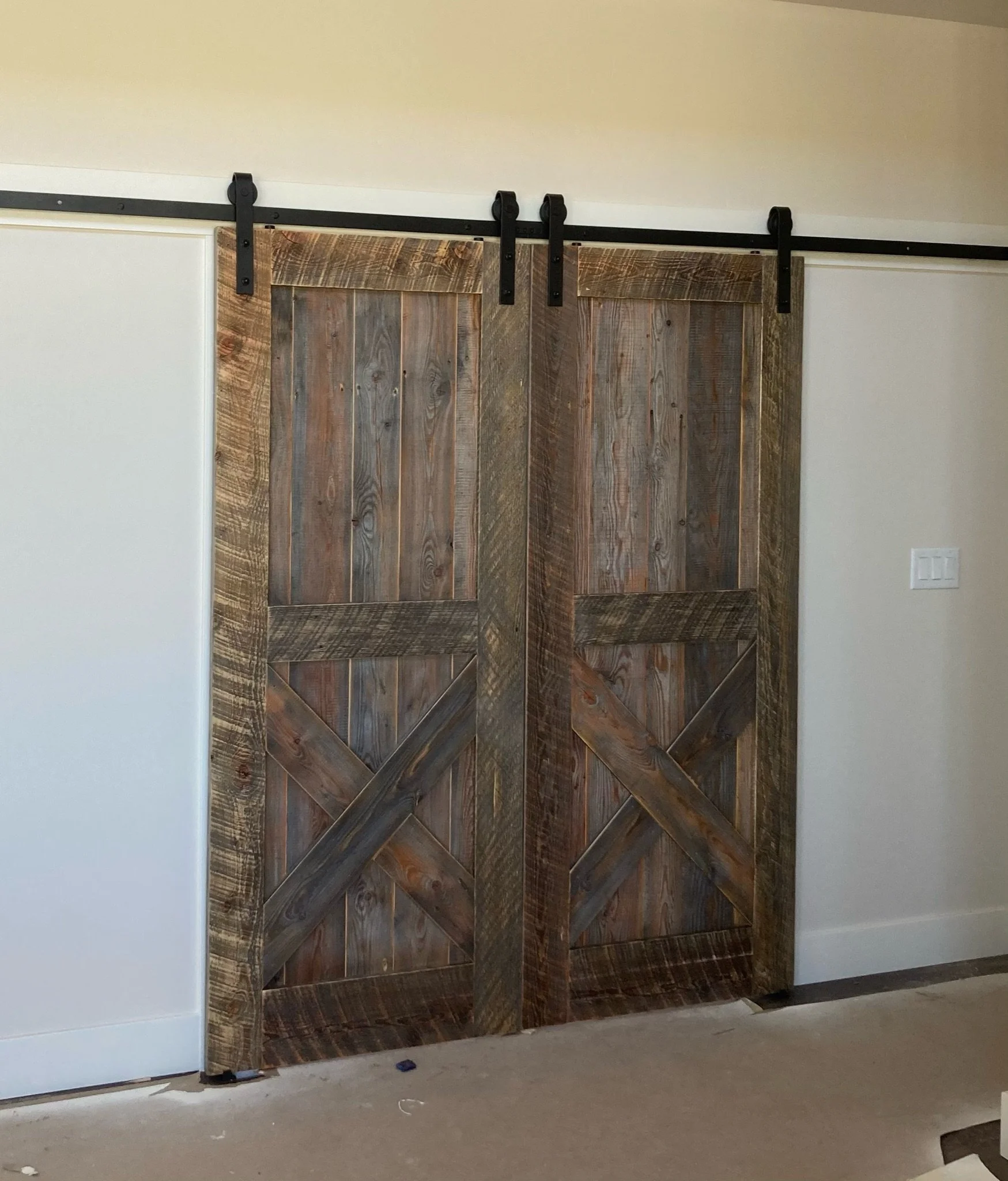 A wooden barn door in a dark stain with black metal roller hardware, partially open in an indoor setting with white walls and a white electrical outlet.