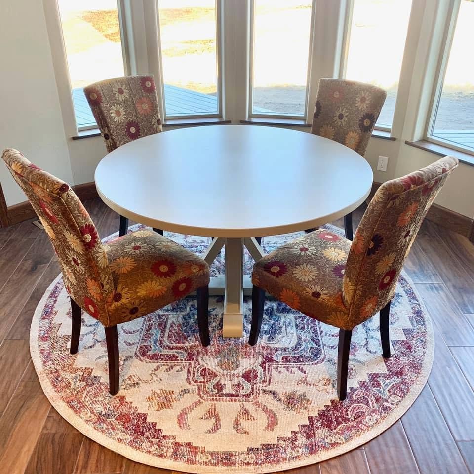 A round dining table with four upholstered chairs featuring floral patterns, set on a circular rug inside a room with large windows showing an outdoor patio.