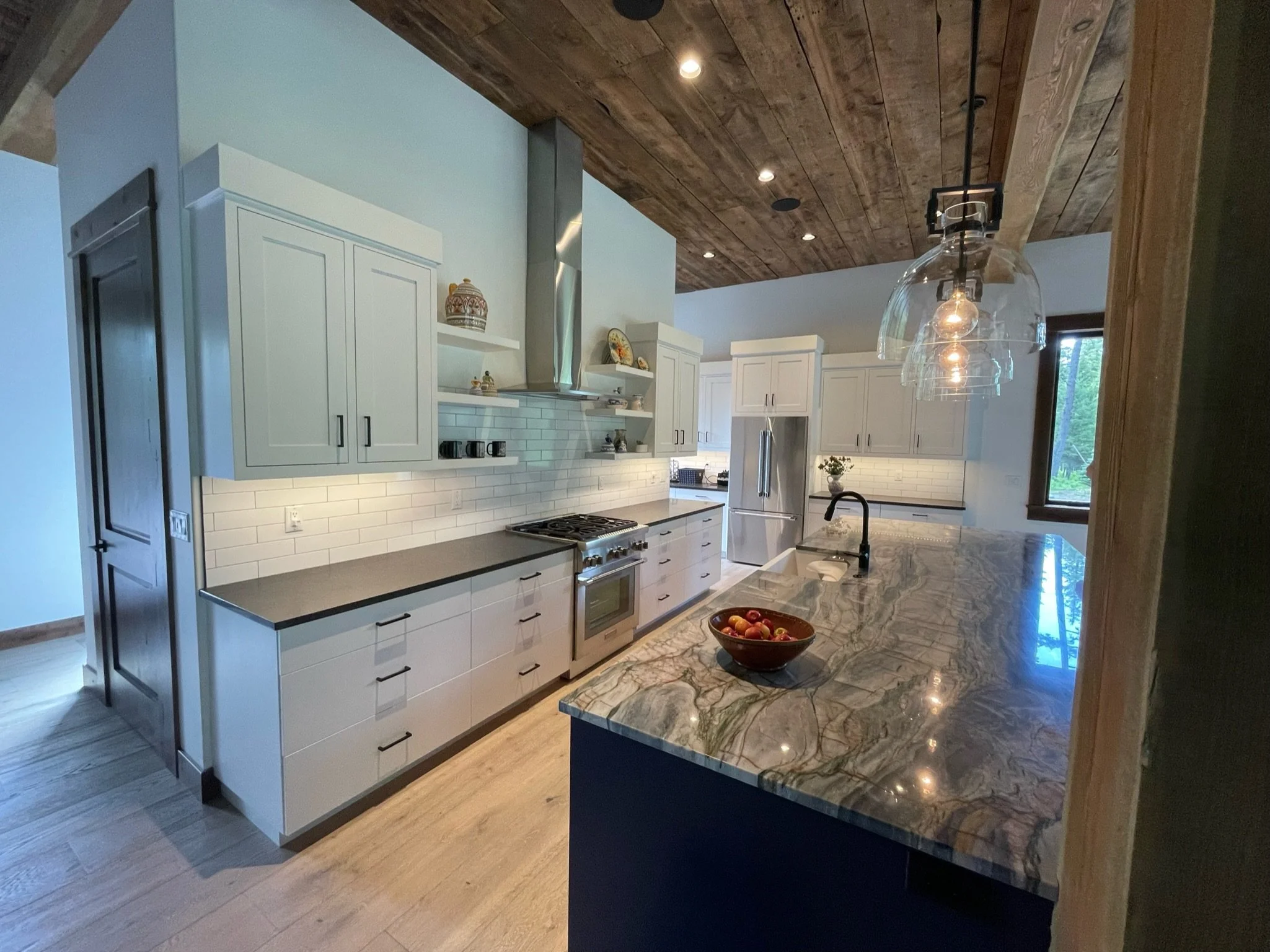 Modern kitchen with white cabinets, a marble countertop, and a dark wood vaulted ceiling. Pendant lights hang above the island, and there are stainless steel appliances including a refrigerator and stove. A bowl of apples is on the island, and a wind