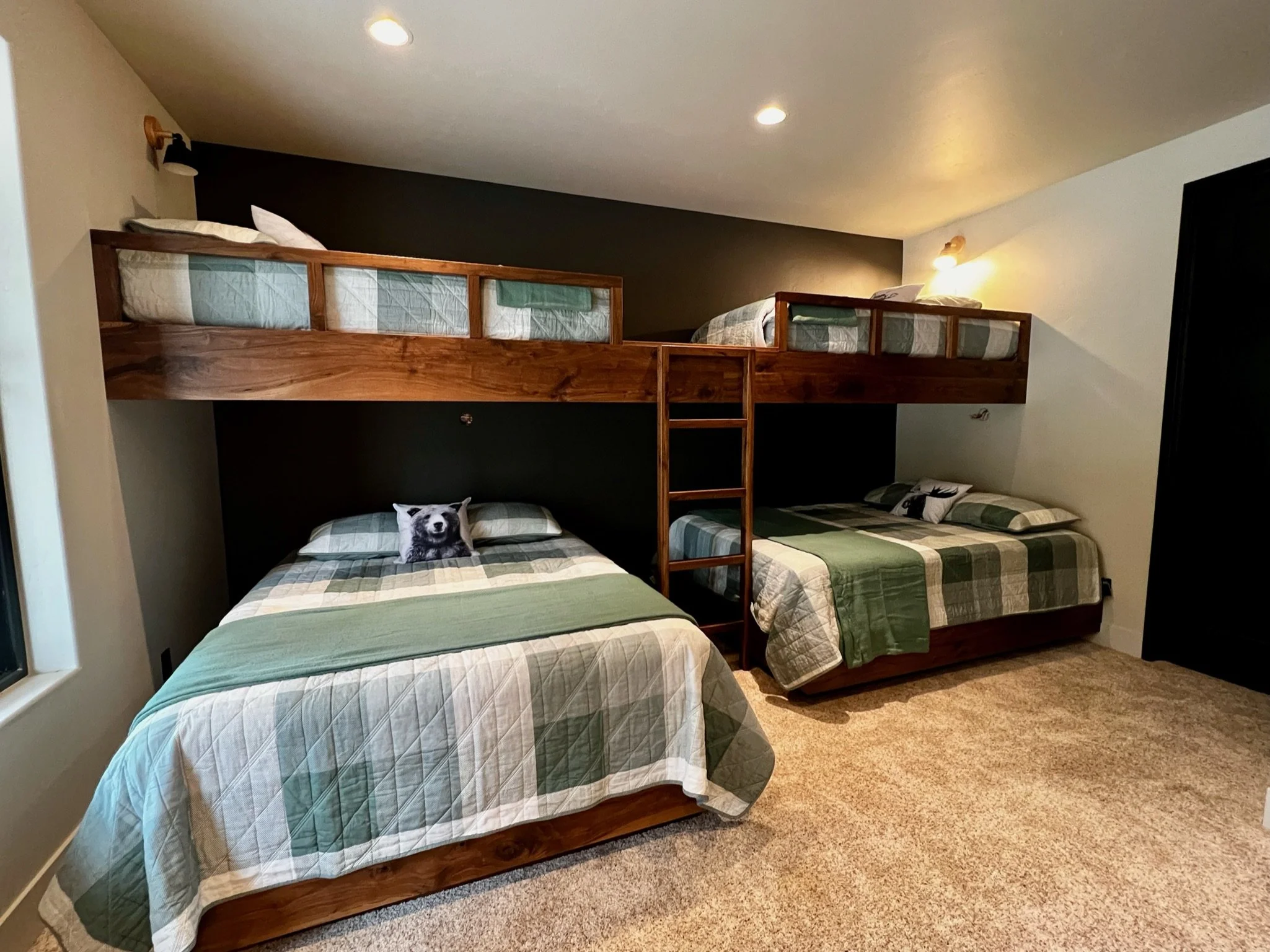A bedroom with three beds: two against the wall, one bunk bed with a ladder, and a dog-themed pillow on the bottom bed, with a wall-mounted light and ceiling lights.