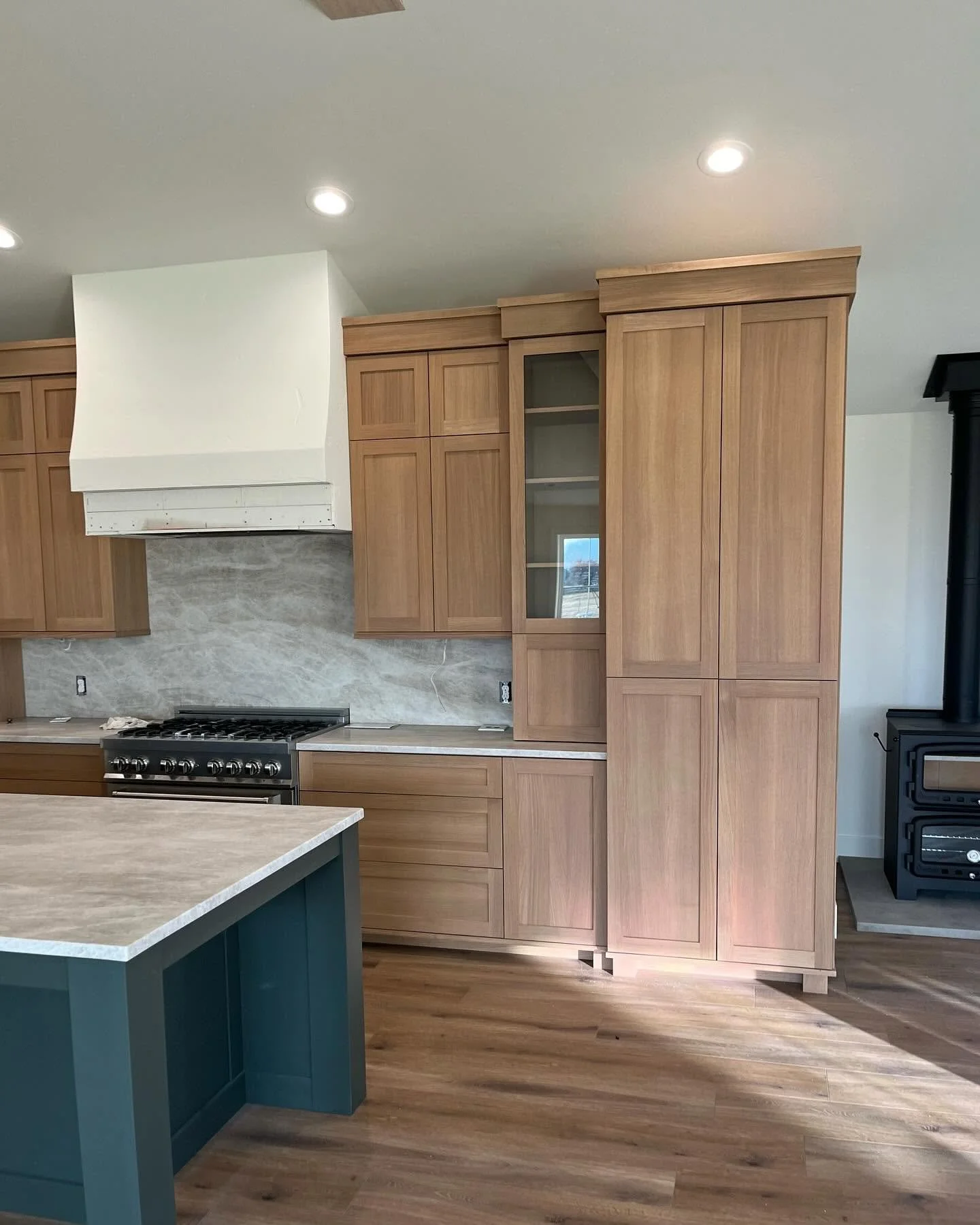 Modern kitchen with wooden cabinets, a white range hood, and a gray marble backsplash. An island with a blue base and beige countertop is visible. A stove and oven are underneath the range hood, and there is a black stove at the side.
