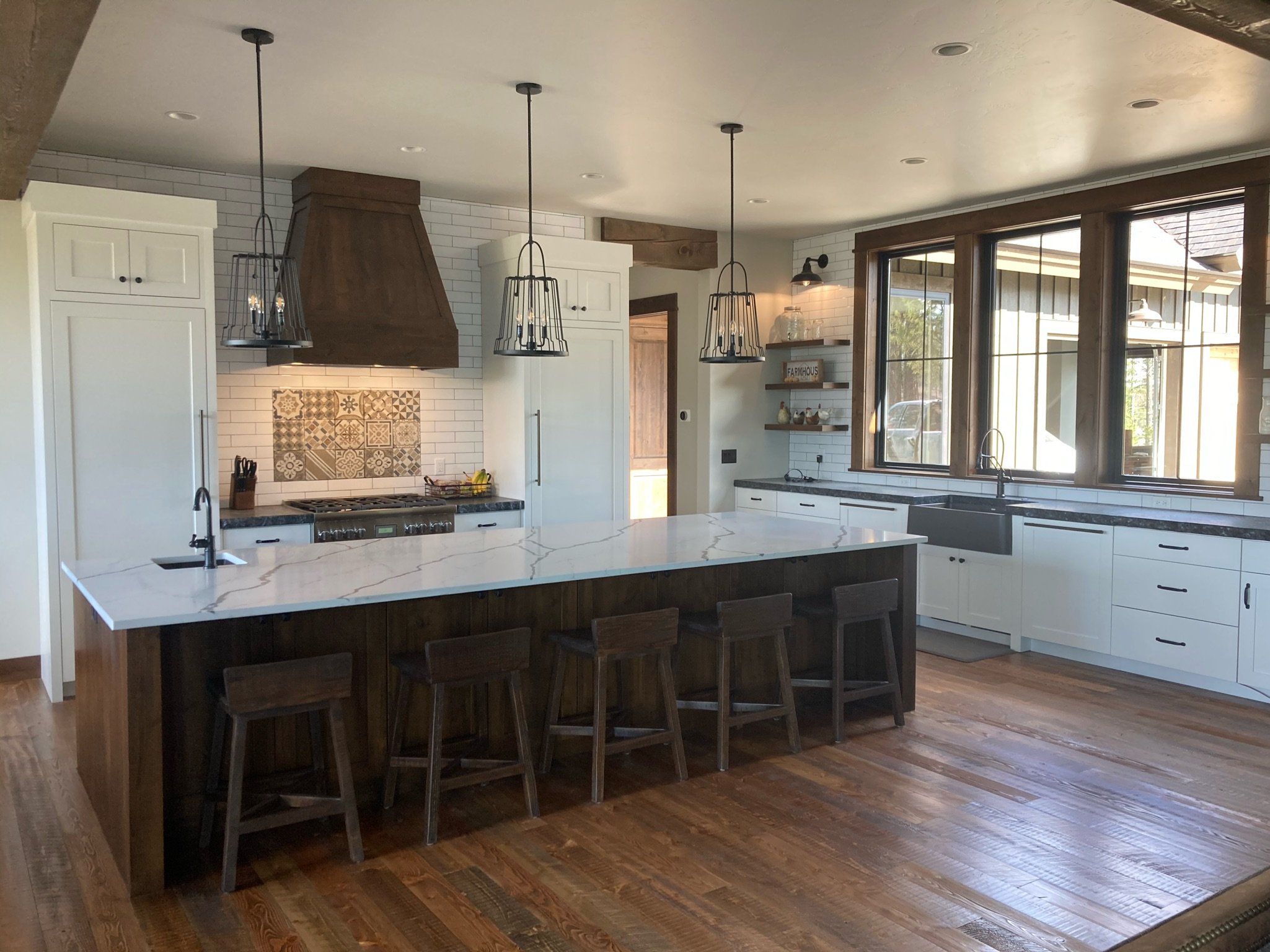 Modern kitchen with white cabinets, a large marble island, black pendant lights, wooden floor, large windows, and a rustic range hood.