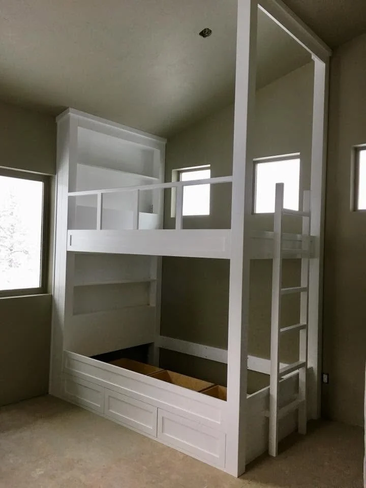 White custom-built loft bed with a staircase, photo taken inside a room with windows and beige carpet.