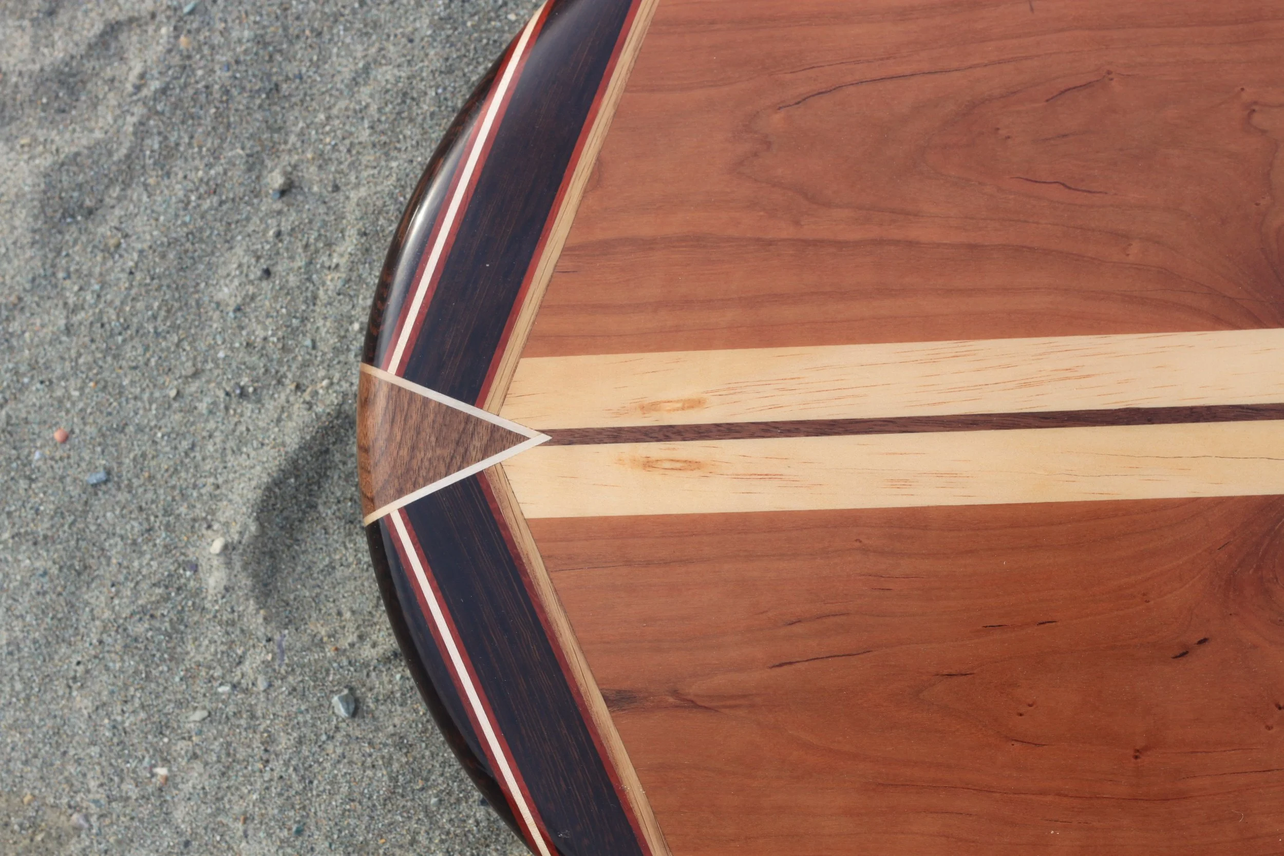 A close-up of the edge of a wooden table with various types of wood and a dark, shiny trim, placed on a gravel surface.