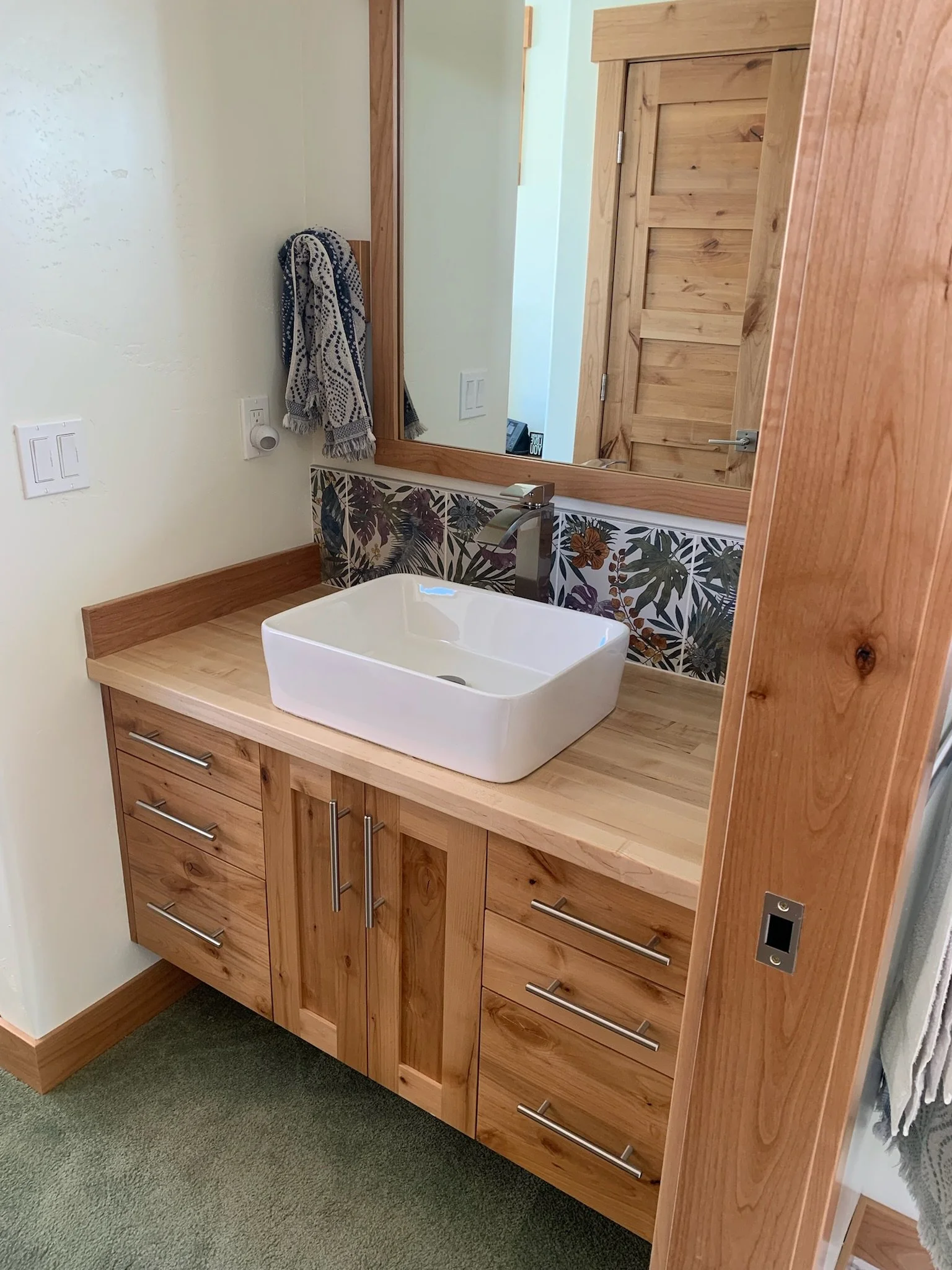 Wooden bathroom vanity with a rectangular white vessel sink, large mirror, and a towel hanging on the wall. The vanity has multiple drawers and cabinets with metallic handles. The wall behind has decorative tile with a botanical pattern.
