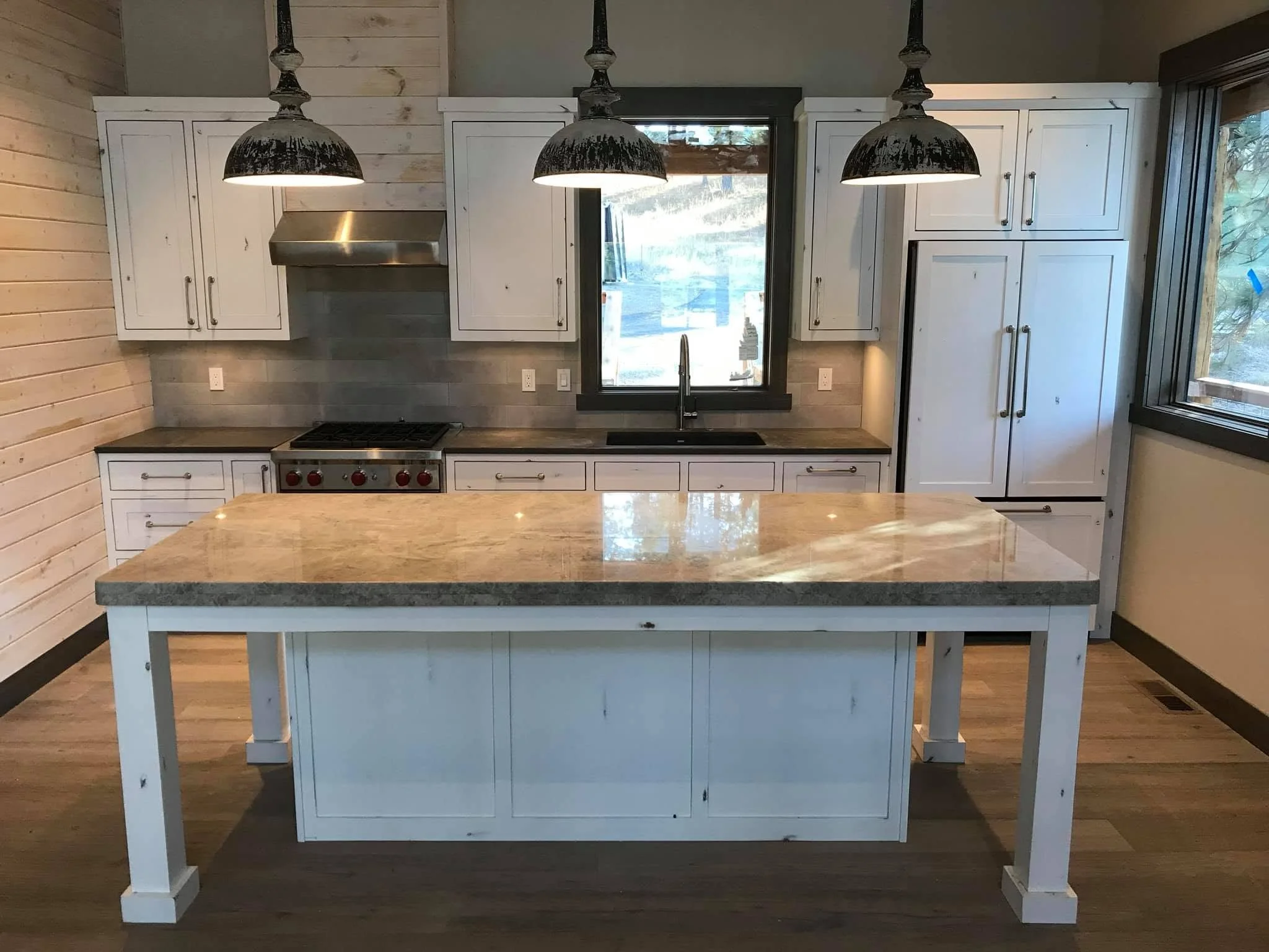 A kitchen with white cabinets, a black-framed window, a kitchen island with a beige marble top, and three black pendant lights hanging above the island.