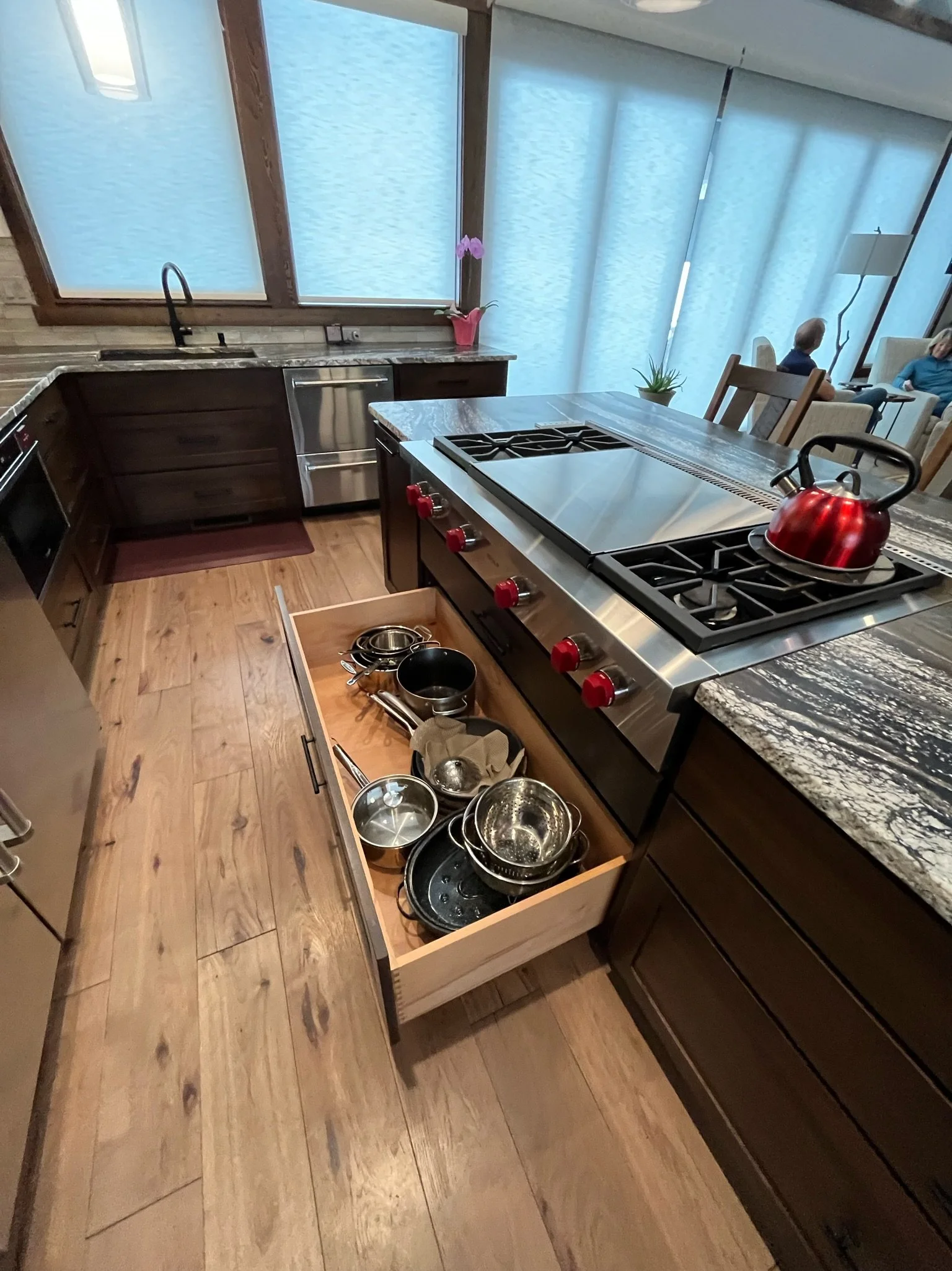 A kitchen with a granite countertop, wooden cabinets, stainless steel appliances, and an open drawer filled with pots, pans, and utensils. In the background, there is a window with frosted glass, a potted plant with pink flowers, and a dining area wi