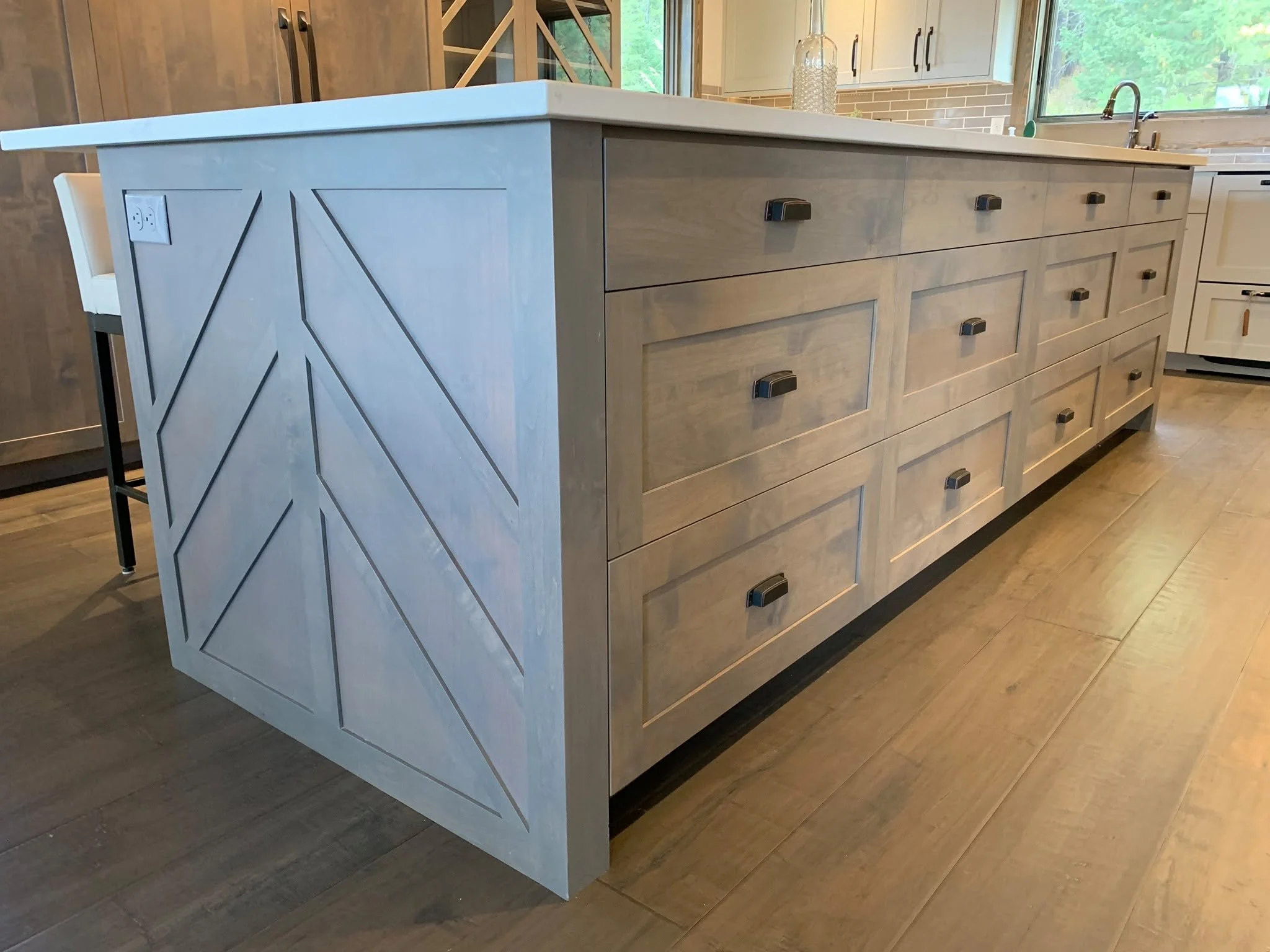 A kitchen island with a light wood finish, featuring multiple drawers with black handles, situated in a modern kitchen with large windows and a brick backsplash.