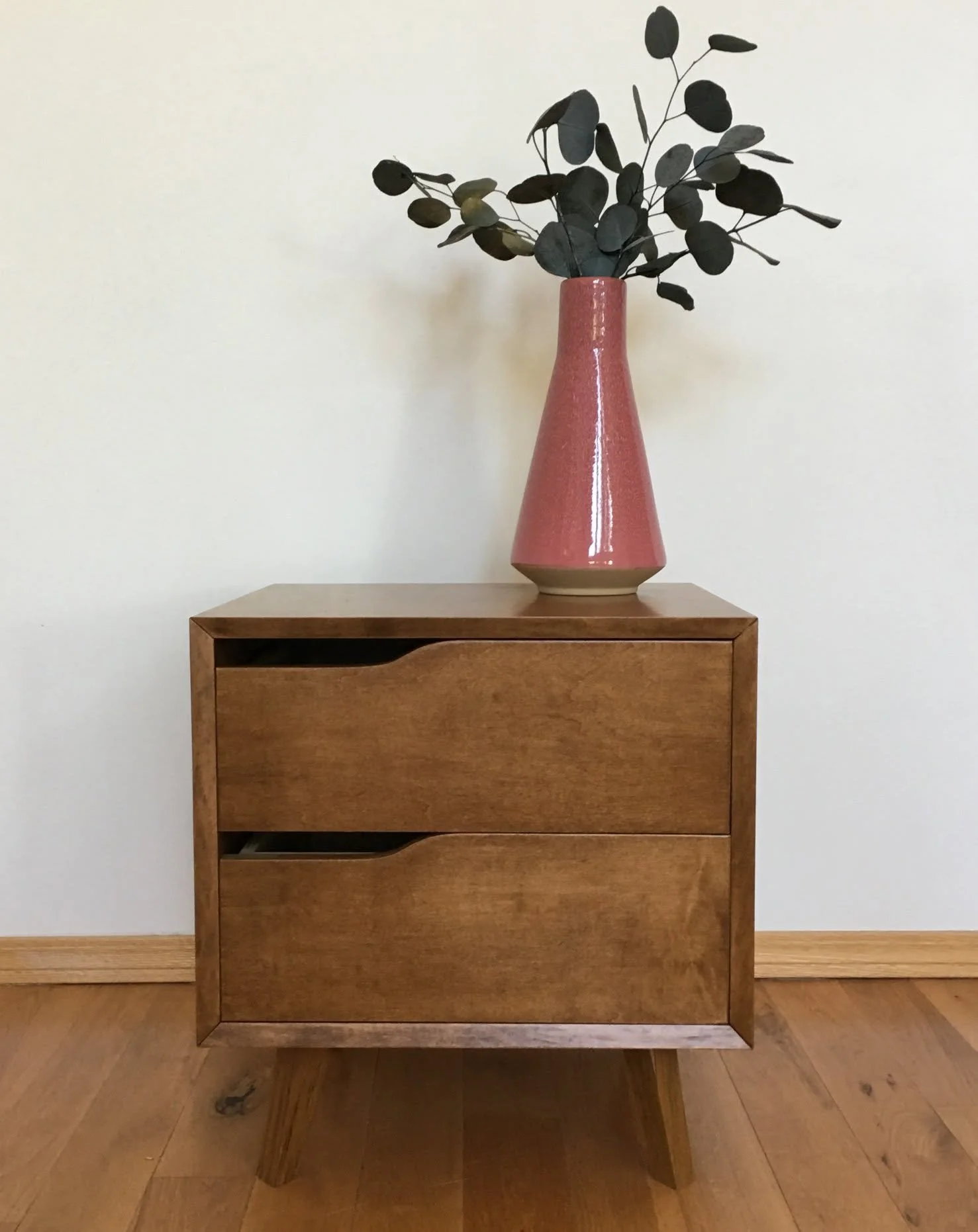 A pink ceramic vase with dark green eucalyptus leaves on a wooden table against a plain white wall.