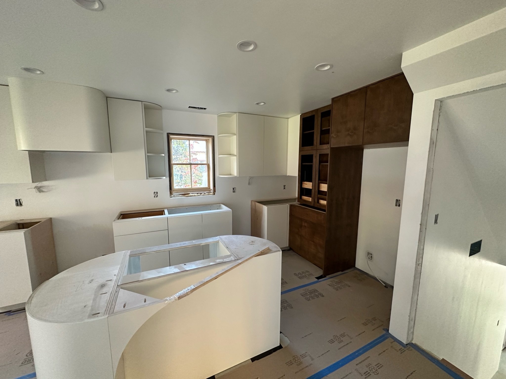 Interior view of a kitchen under construction with white cabinets, a curved island, and a wooden wall cabinet, with a window showing trees outside.