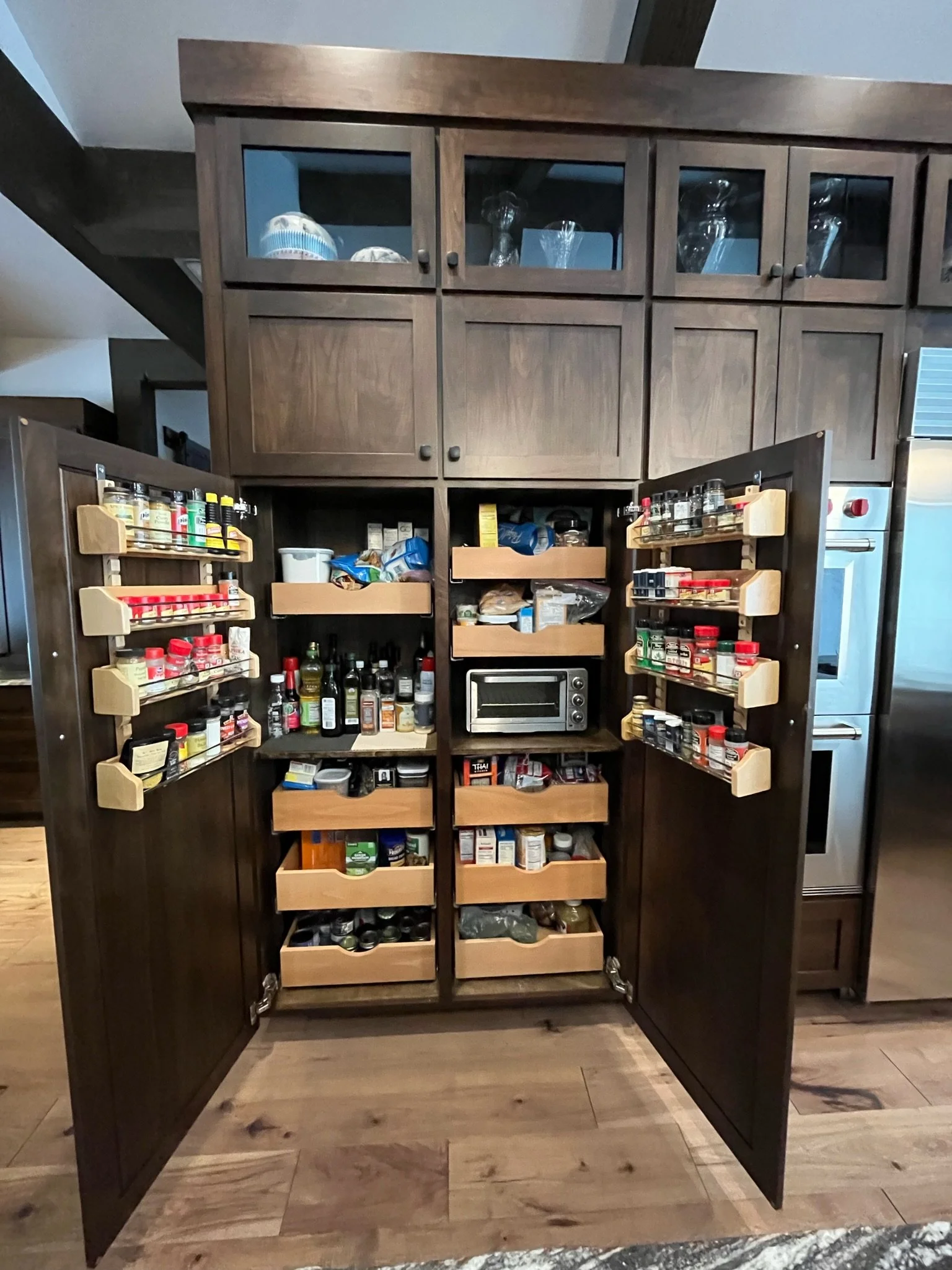 A dark wooden kitchen cabinet with glass doors on the upper section, containing glassware, and open drawers and shelves below filled with spices, condiments, and food items. A small oven or toaster oven is also in the open lower section.