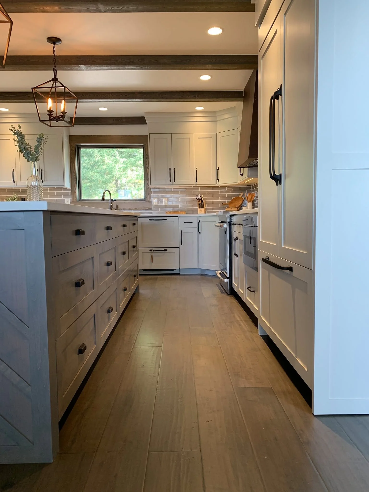 A modern kitchen with white cabinets, dark handles, a window above the sink with a view of trees, beige tile backsplash, kitchen island with drawers, and stainless steel appliances. There is a black metal light fixture hanging from the ceiling and re