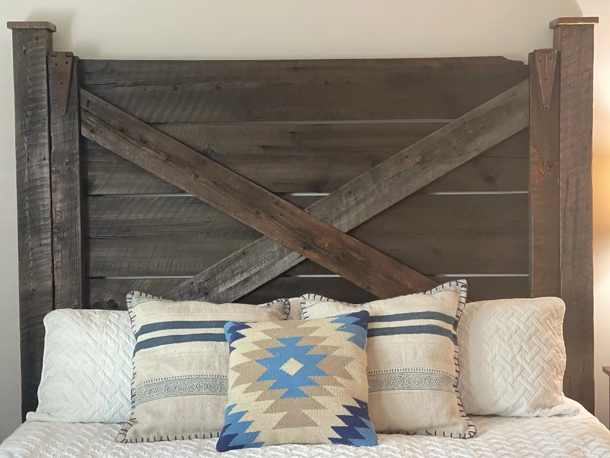 A rustic wooden headboard with diagonal supports behind a bed with pillows that have southwestern-style patterns in blue, beige, and black. There is a quilted bedspread in white.