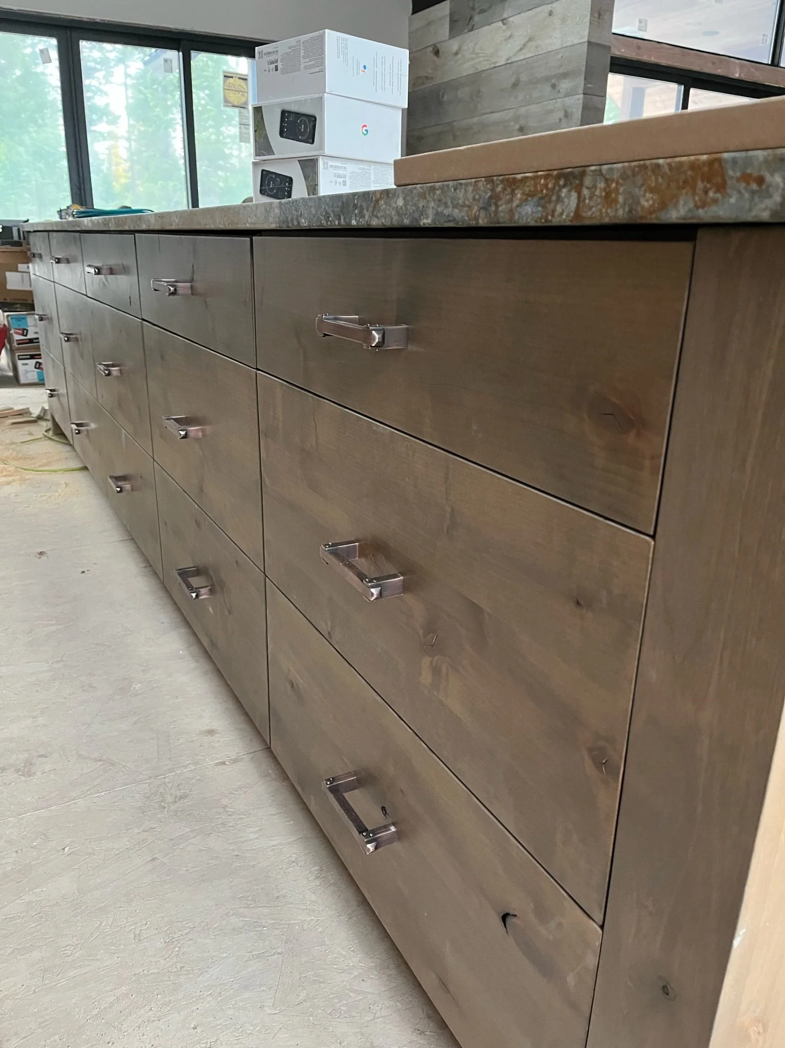 A wooden kitchen cabinet with multiple drawers and metal handles, situated beneath a countertop, with a window and some boxes in the background.