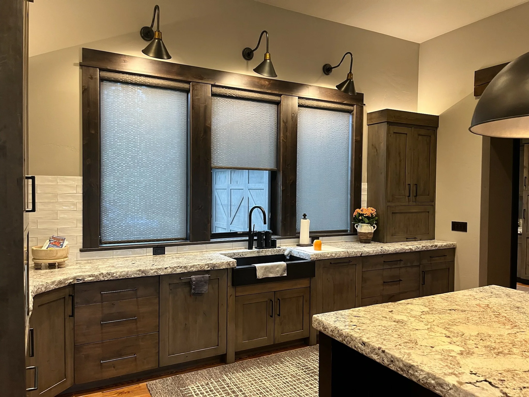 A kitchen with dark wooden cabinets and a speckled granite countertop. Above the window, there are three black wall-mounted lamps with gold accents. The window has frosted glass panels with a textured pattern. On the countertop, there is a white pape