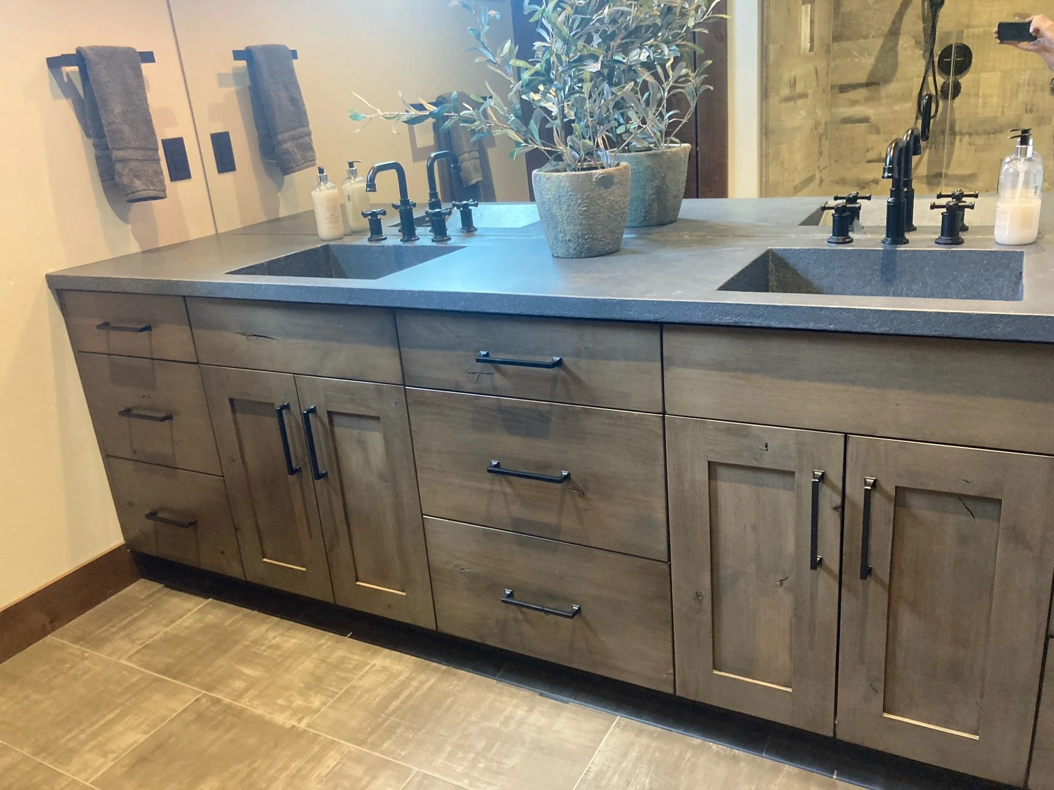 Bathroom vanity with two sinks, black fixtures, wooden cabinet, potted plant in the center, hand towels on the wall, soap dispensers, and a mirror reflecting part of the bathroom.