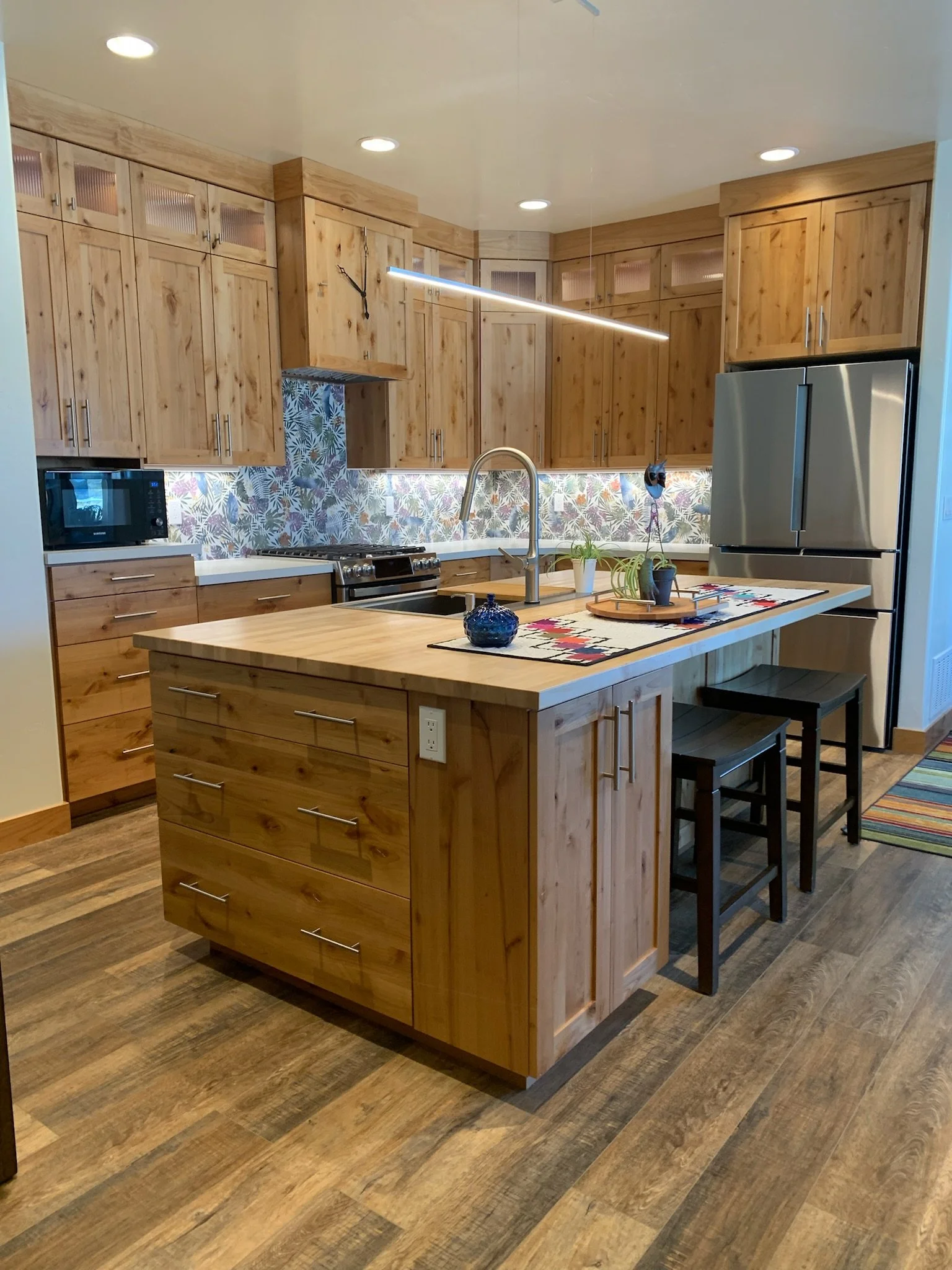 A modern kitchen with wooden cabinets, a kitchen island, stainless steel refrigerator, black stools, and a colorful rug. There are decorative plants and a wall clock on the wall.