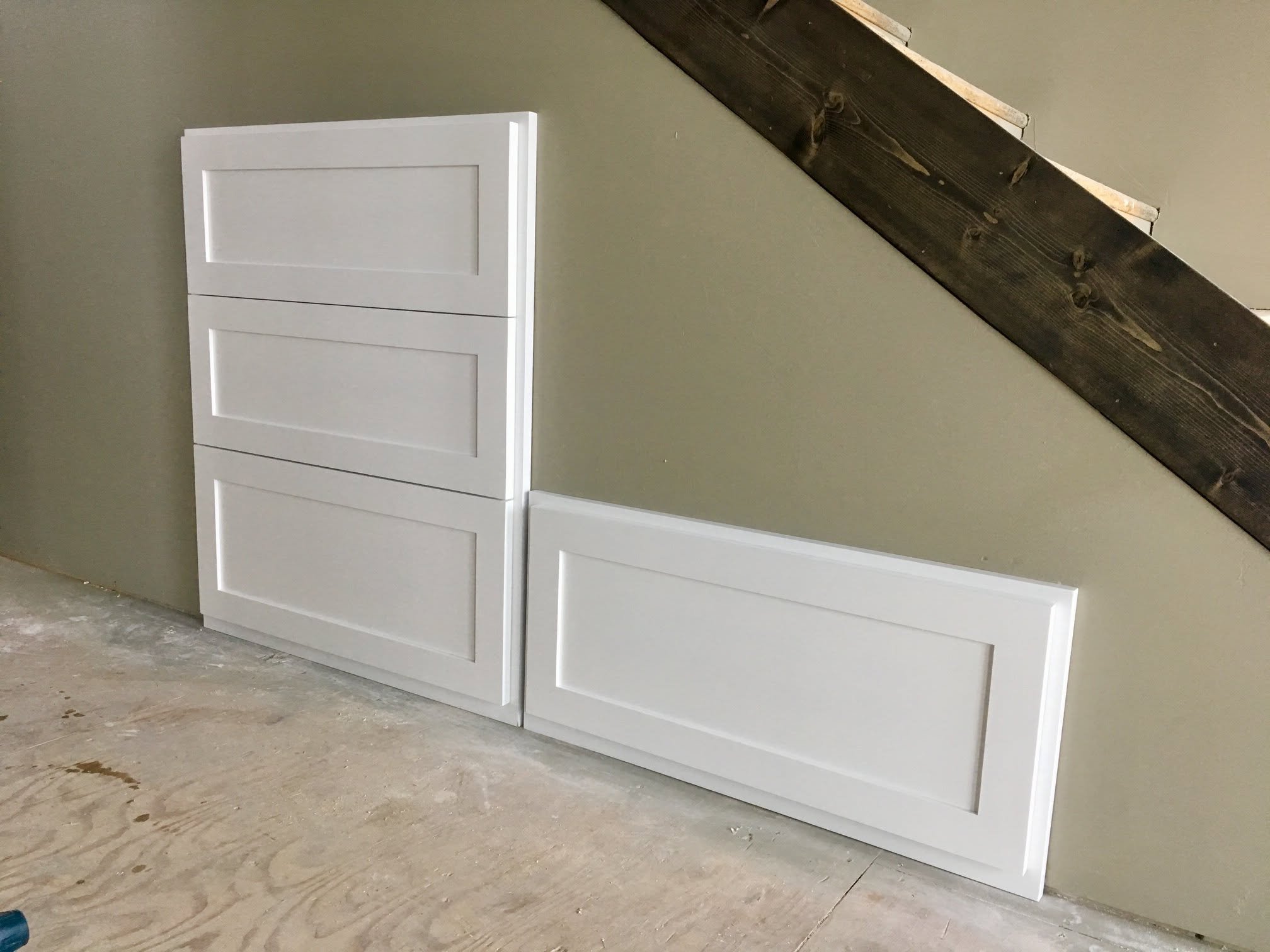 White cabinet or drawer set positioned against a gray wall, with a dark wooden beam above on the wall. The floor appears to be unfinished concrete with visible wood grain and construction debris.