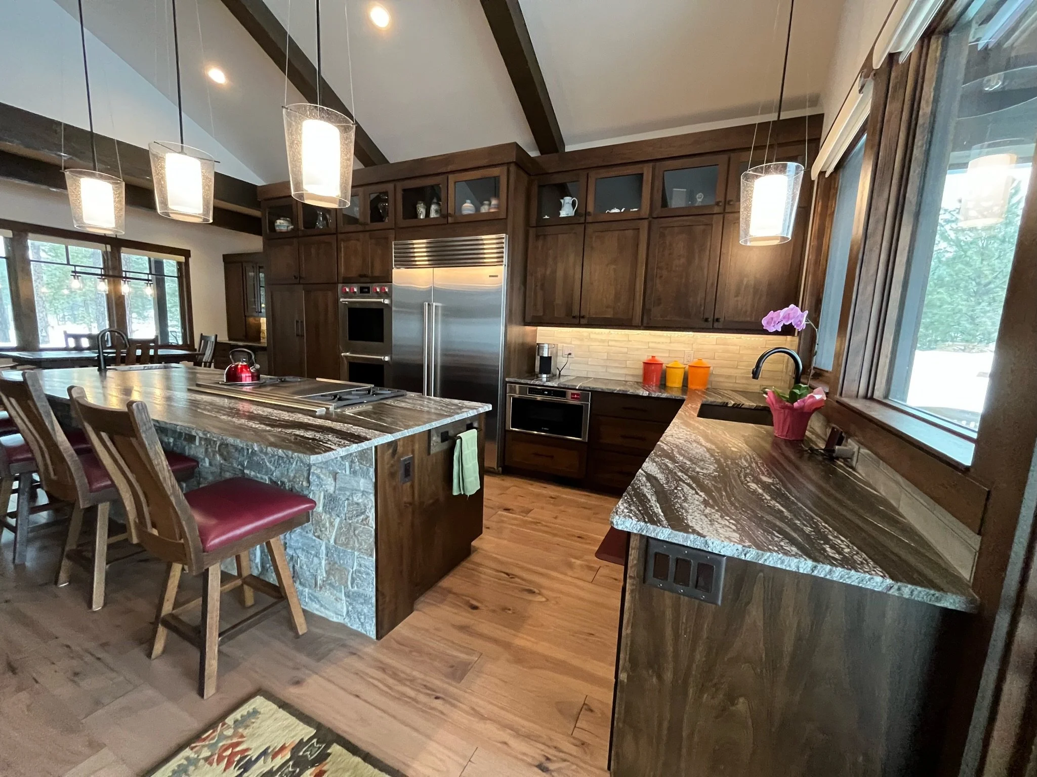 A modern kitchen with wooden cabinets, a granite island, and stainless steel appliances. Pendant lights hang above the island, and large windows let in natural light. There is a orchid plant on the counter and a colorful rug on the wooden floor.