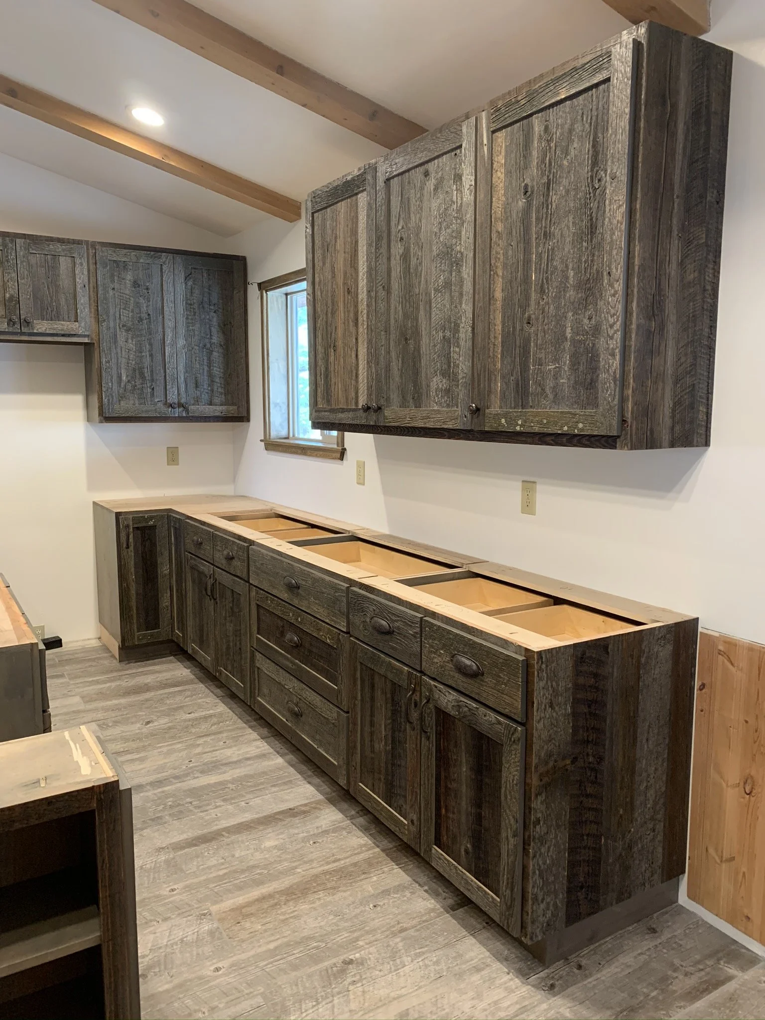 Several dark wood kitchen cabinets are being installed, with some of the doors and drawers missing. The cabinets are beneath a window, and the room features wood ceiling beams and gray wood flooring.