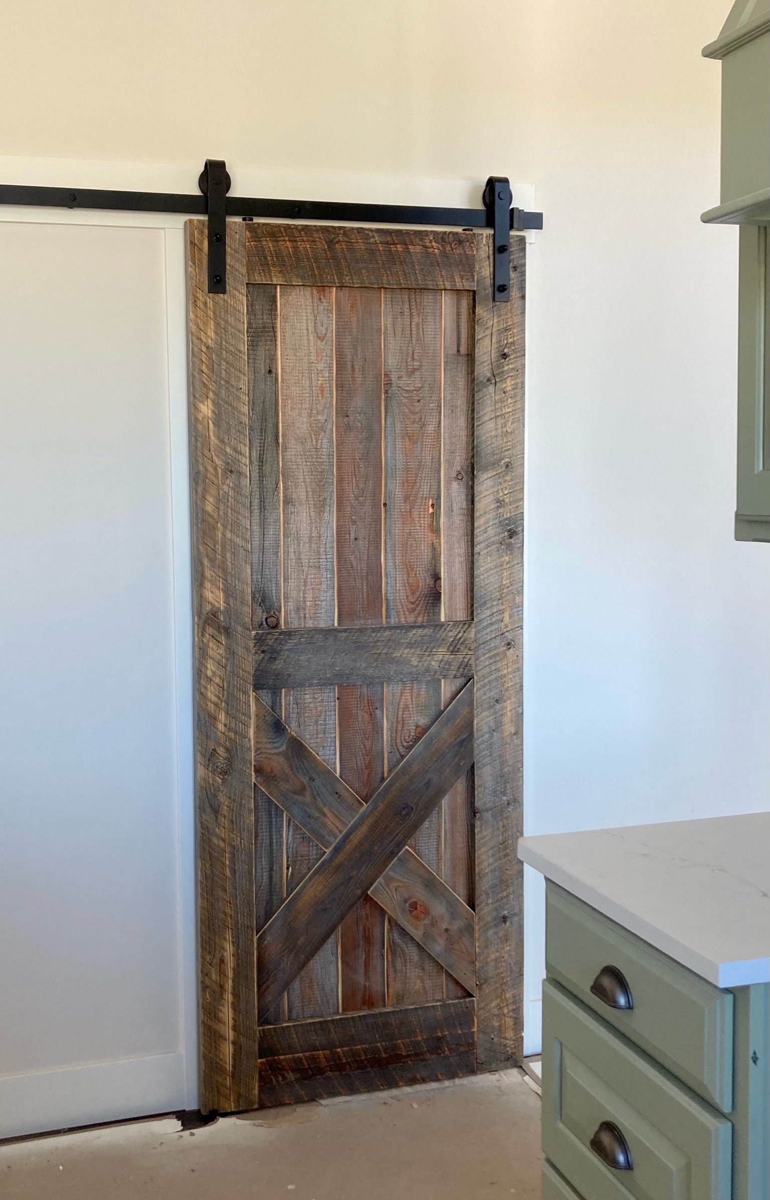 A wooden sliding barn door installed on a black metal rail in a room with light-colored walls and a green dresser with drawers.