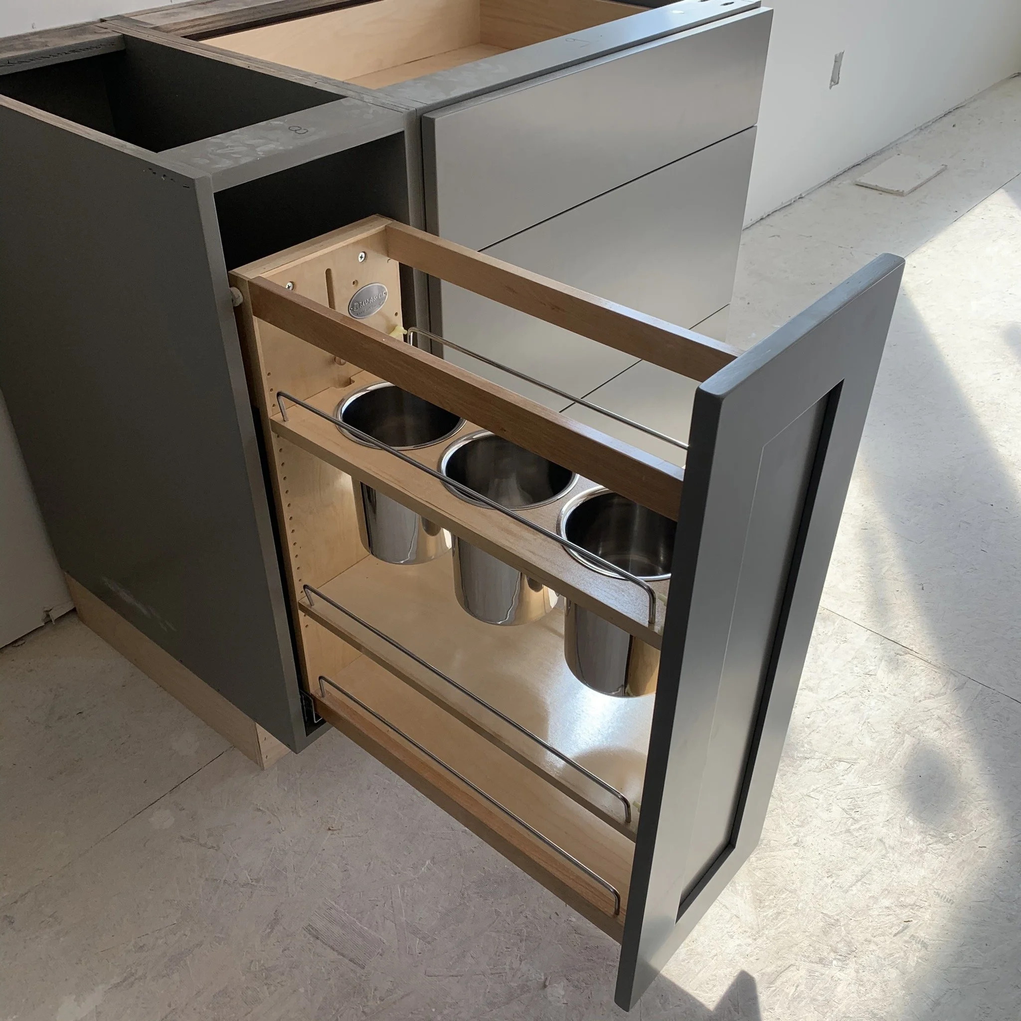 Unfinished kitchen cabinet with gray and wooden panels, featuring two open metal bowls at the bottom, set on a concrete floor in an ongoing construction or renovation area.