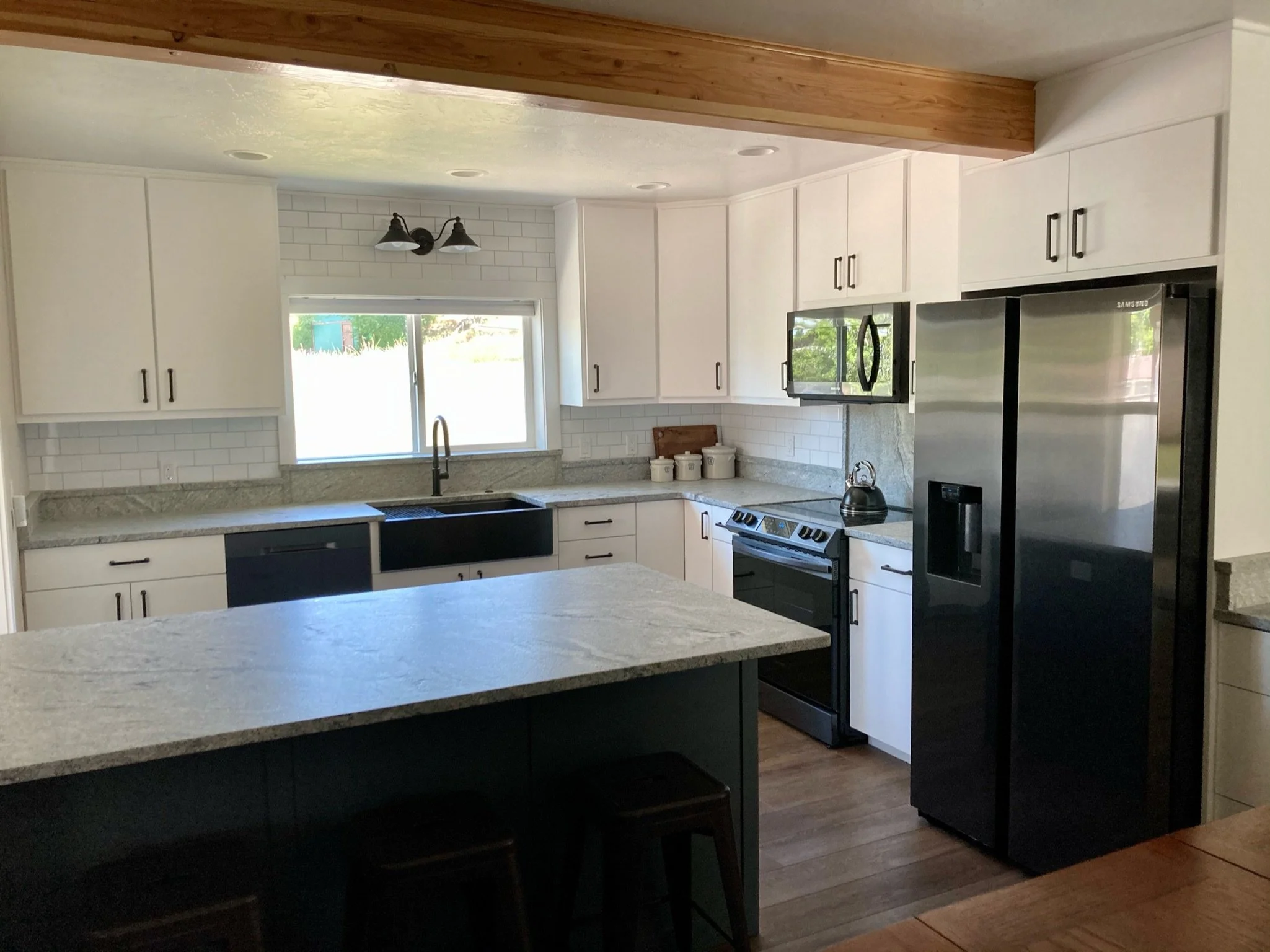 Kitchen with white cabinets, granite countertops, stainless steel refrigerator, black stove, microwave, and black knobs, with a window above the sink and a wooden ceiling beam.