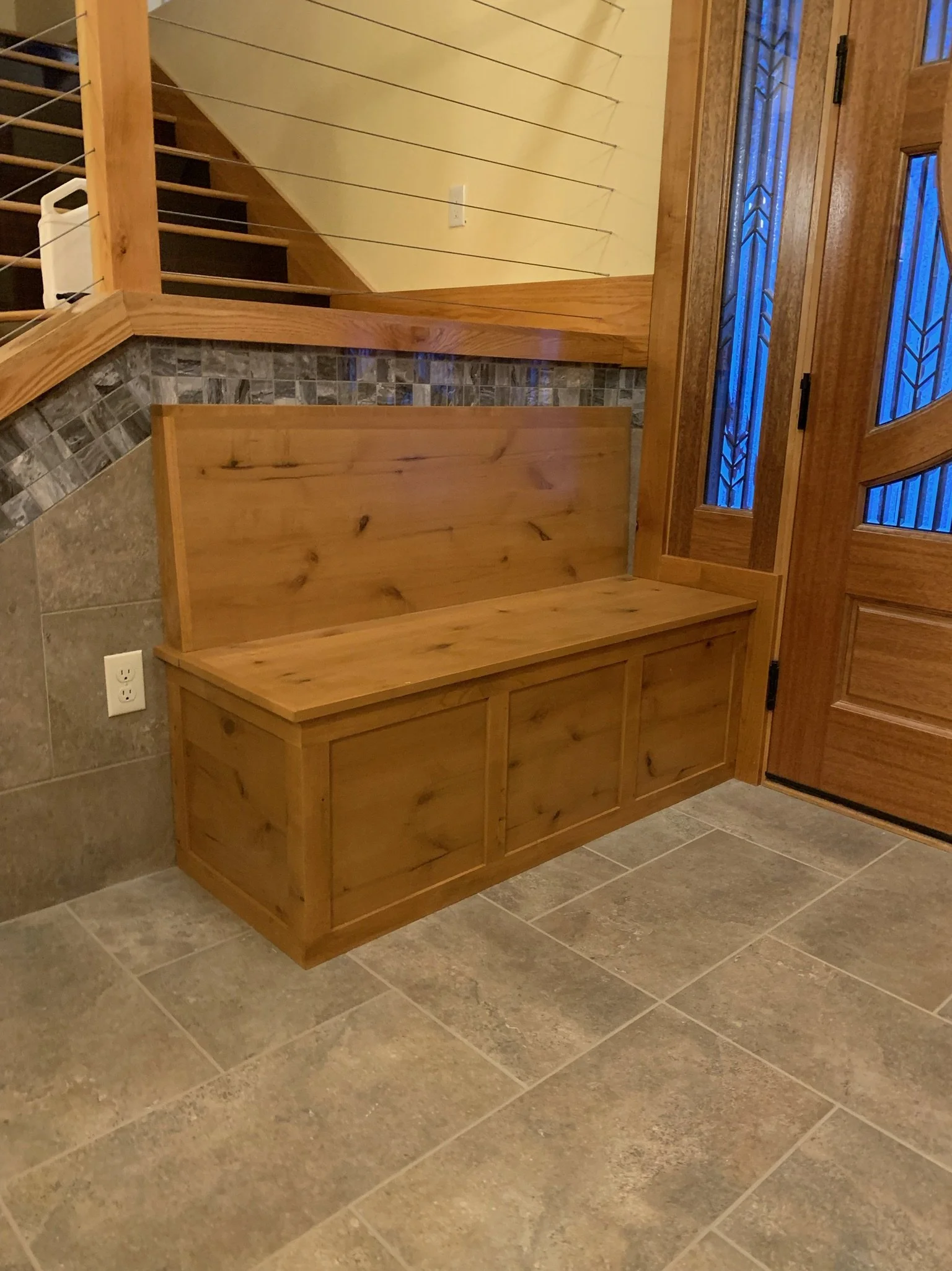 Wooden bench with cabinet doors underneath, against a wall with mixed tile and wood finish, next to a door with decorative glass panels.