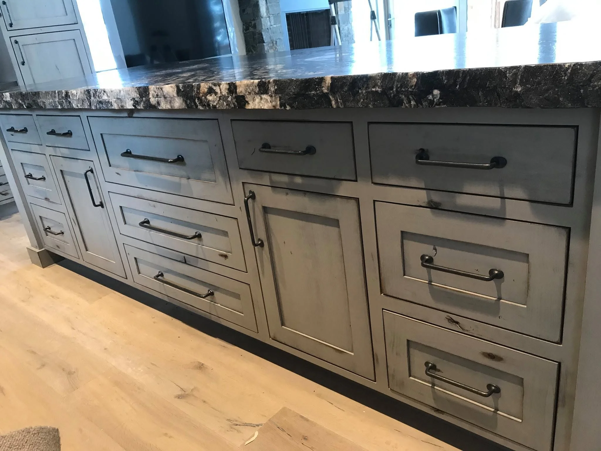 Close-up of a light gray kitchen cabinet with a granite countertop, featuring several drawers and black handles, underneath a reflective surface in a modern kitchen setting.