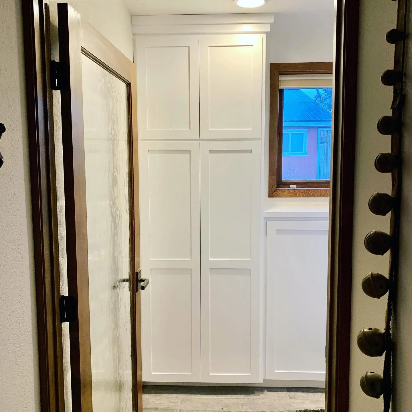 Small room with a white cabinet, a window with wooden trim showing neighboring house, and a door with brown trim reflected in a mirror.