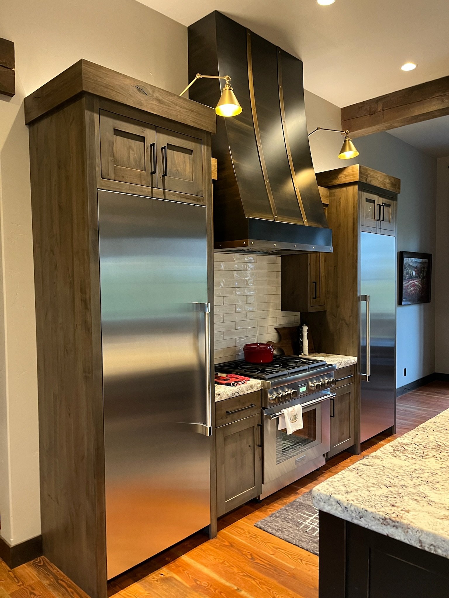 Kitchen with wooden cabinets, stainless steel refrigerator, and a stove with a red pot on top. The ceiling has recessed lighting, and there is granite countertop in the foreground.
