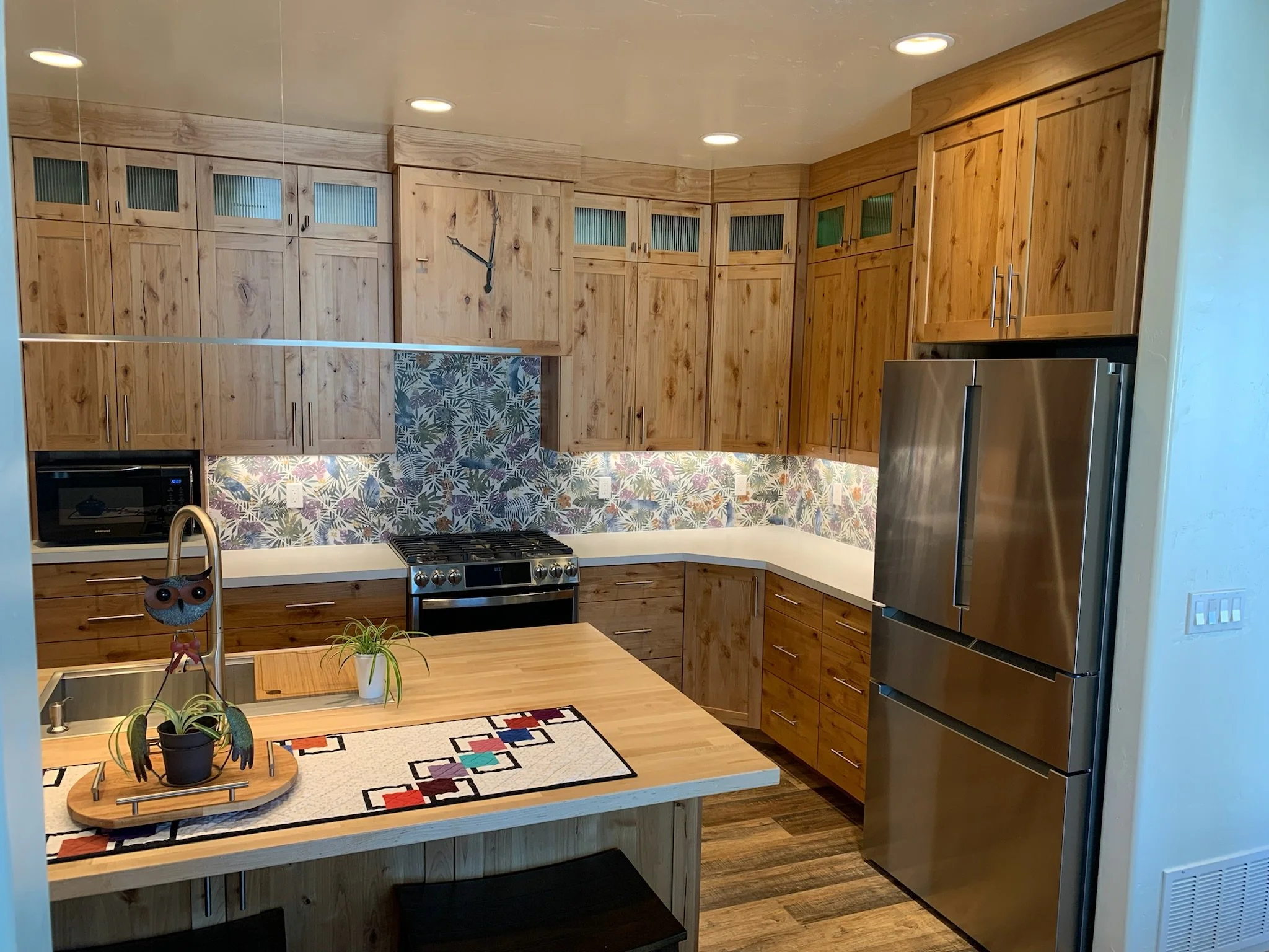 Modern kitchen with wooden cabinets, stainless steel refrigerator, gas stove, decorative tile backsplash, and a countertop with a plant and owl-shaped decor.