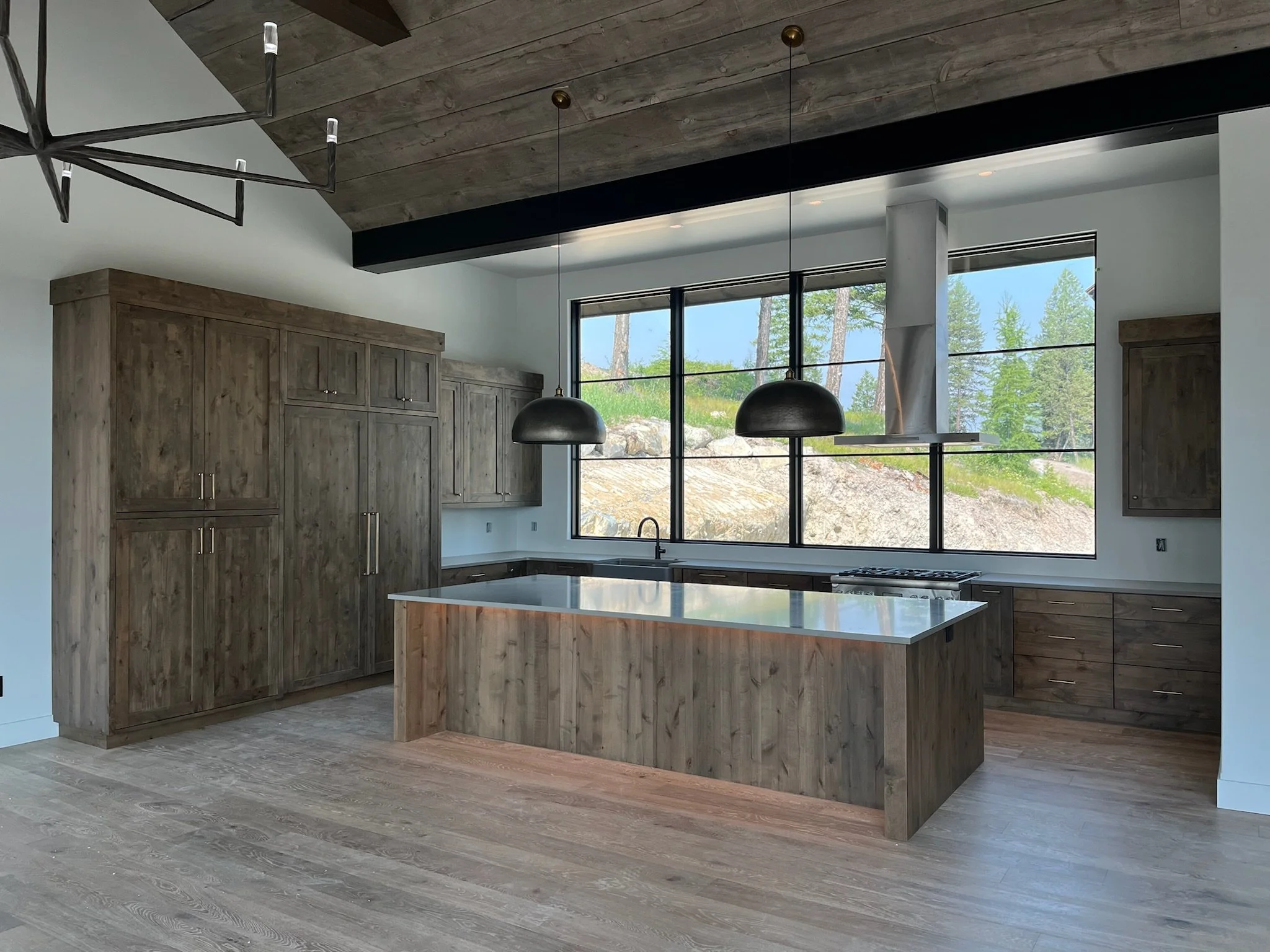 Contemporary kitchen with wooden cabinets, a large island, black pendant lights, large windows showing a green outdoor landscape, and a wooden floor.