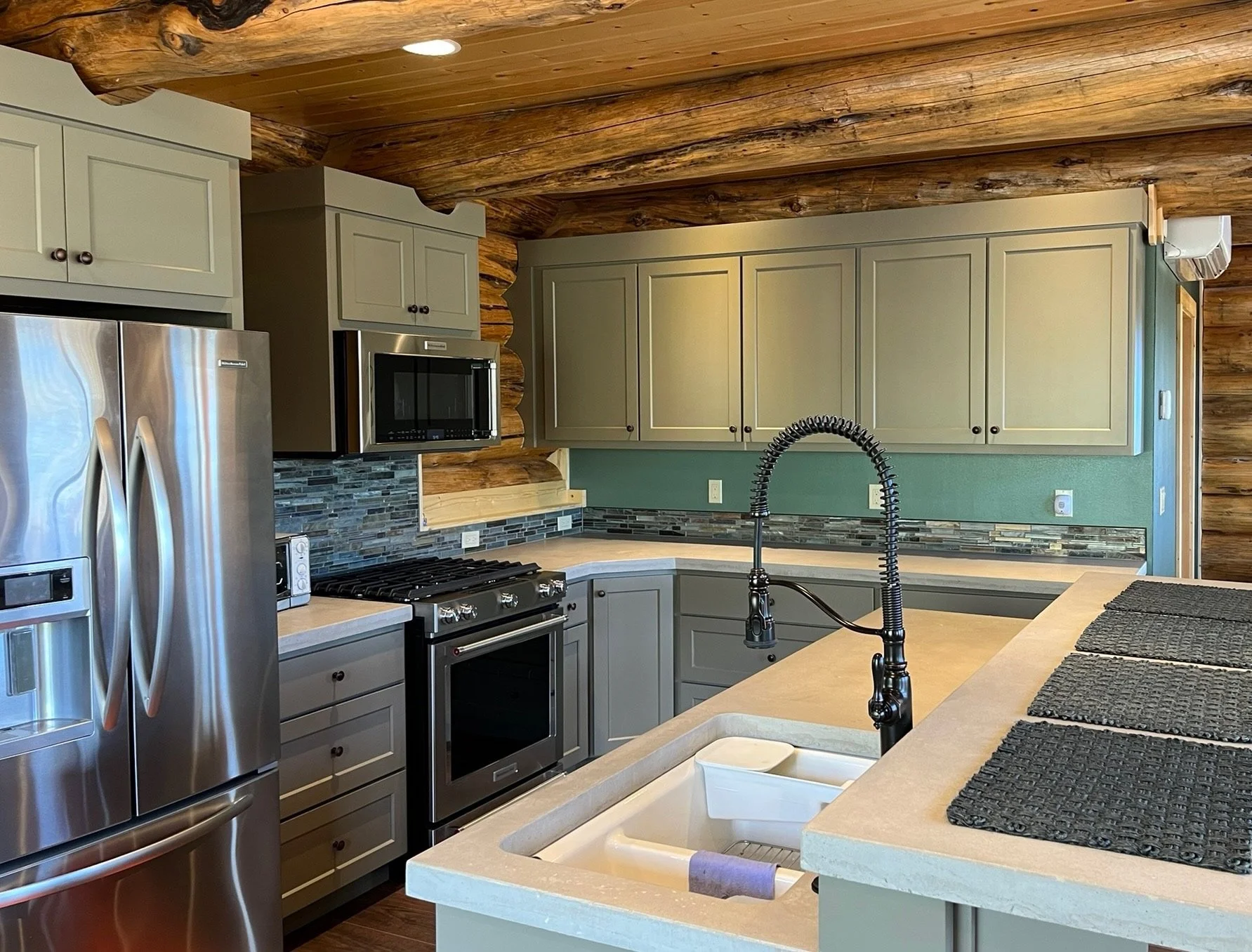 Kitchen with wooden ceiling and gray cabinets, stainless steel refrigerator, black stove, microwave, tiled backsplash, green wall, and a beige countertop with black placemats.