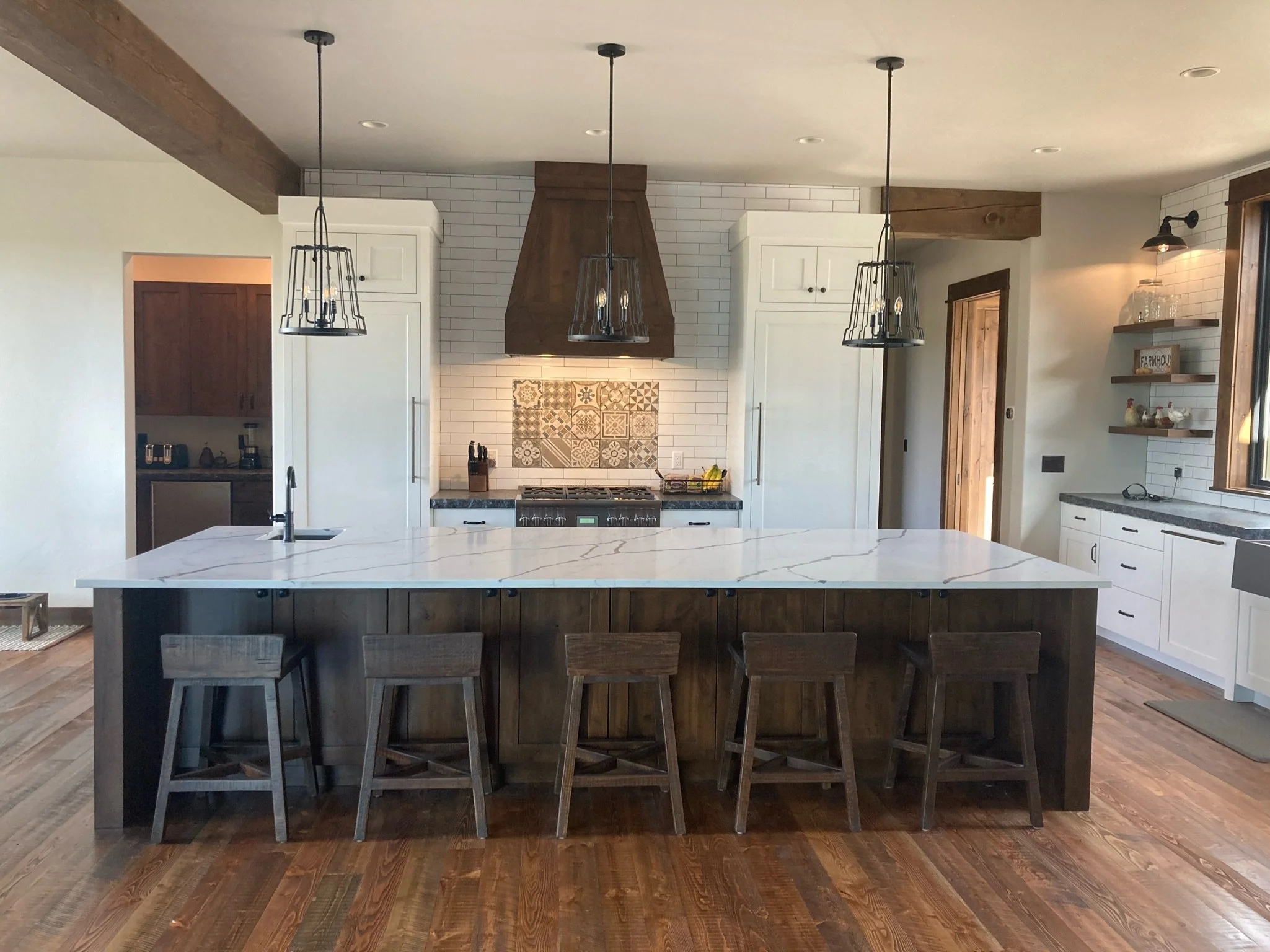 Modern kitchen with white cabinets, a large marble island, black pendant lights, a stove with an overhead wooden range hood, and white subway tile backsplash with decorative tiles in the center.