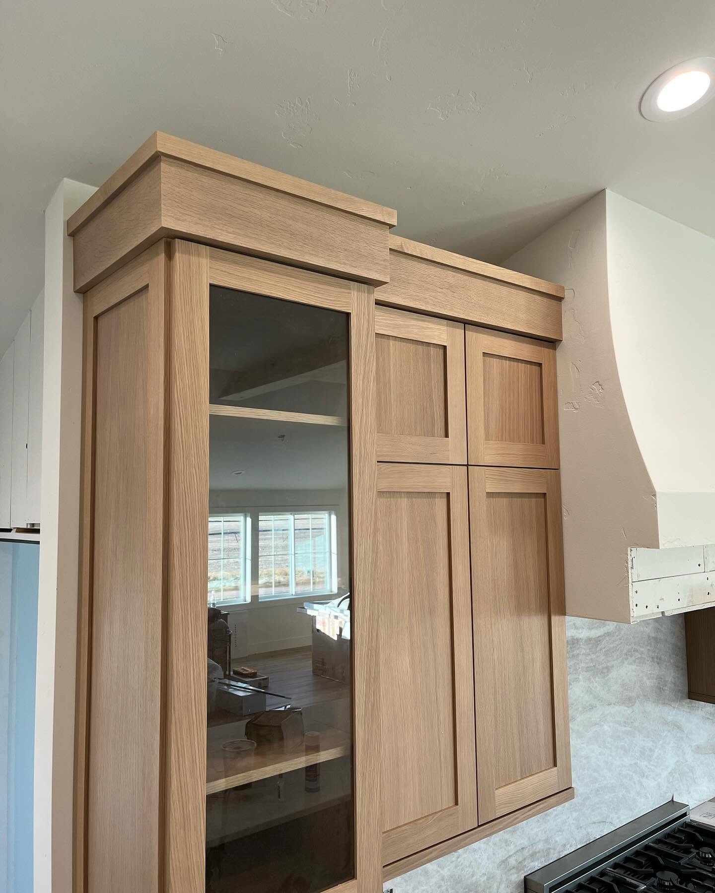 A wooden kitchen cabinet with sliding glass doors and a mirror, situated above a light-colored countertop in a kitchen, with a window and part of a stove visible.