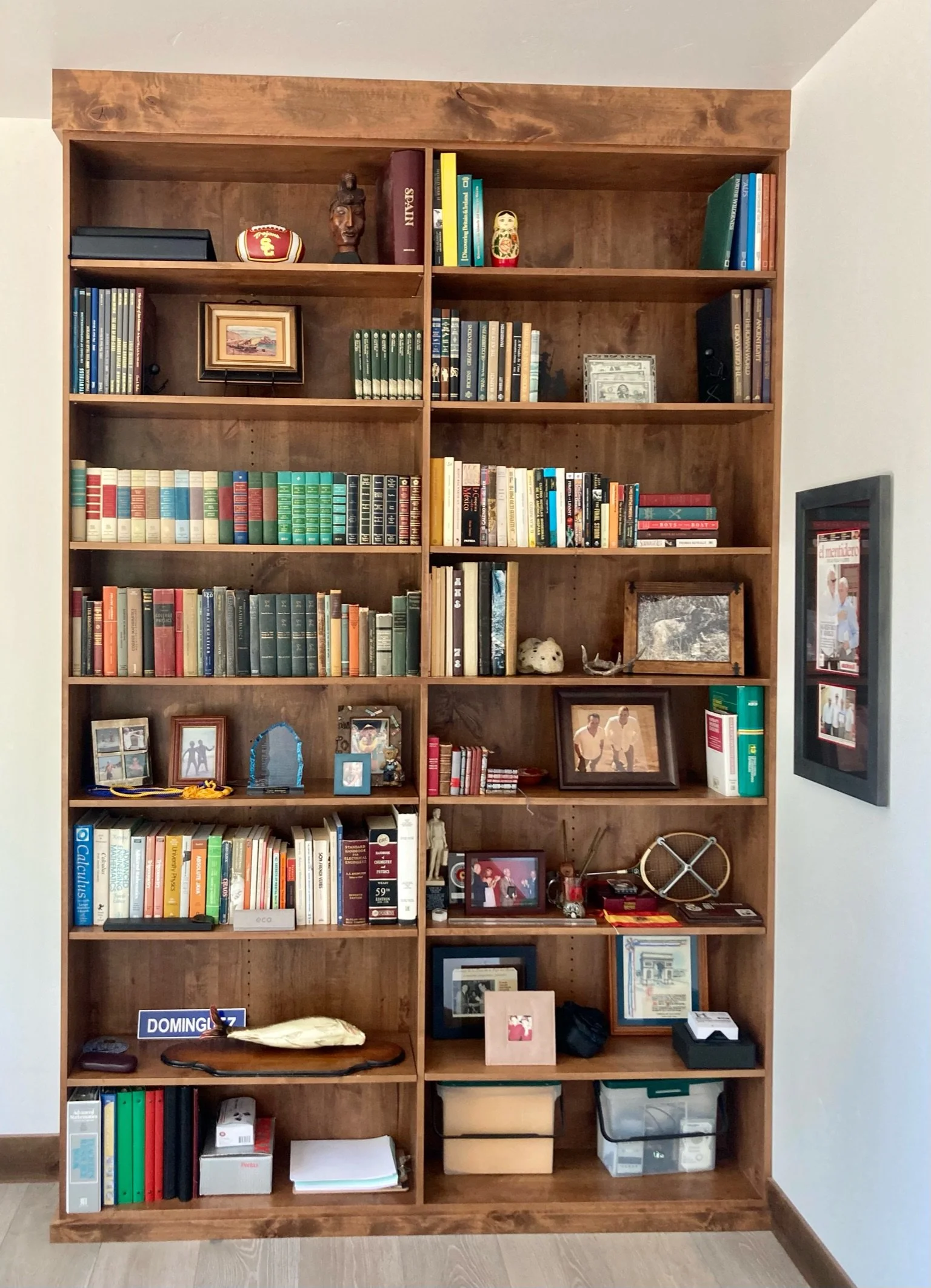 A large wooden bookshelf filled with books, framed photographs, small decorative items, and office supplies in a room with light-colored walls and hardwood flooring.