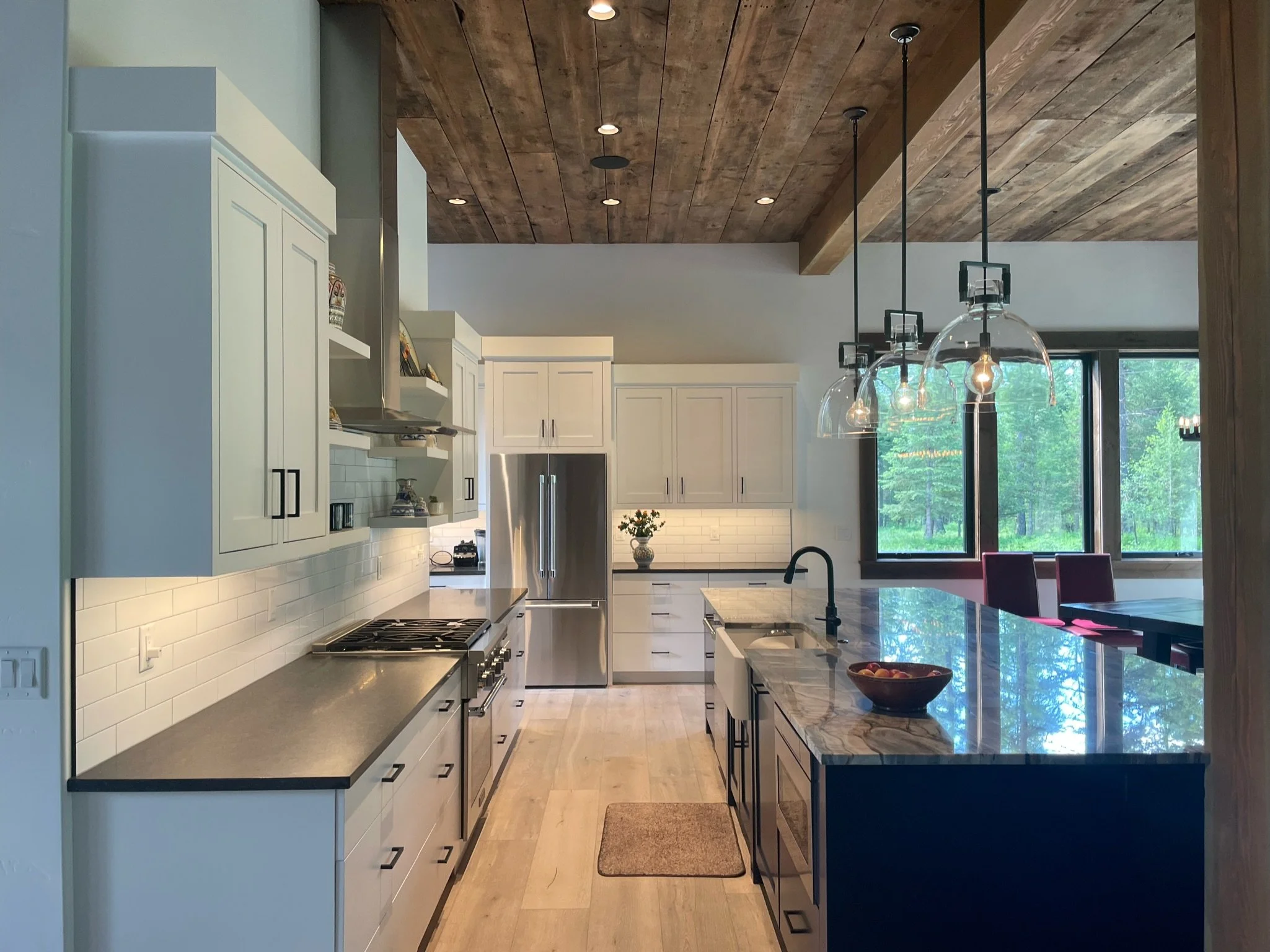 Modern kitchen with white cabinets, gray countertops, a wooden ceiling with recessed lighting, pendant lights over island, stainless steel appliances, a window showing greenery outside, and a small rug on the floor.