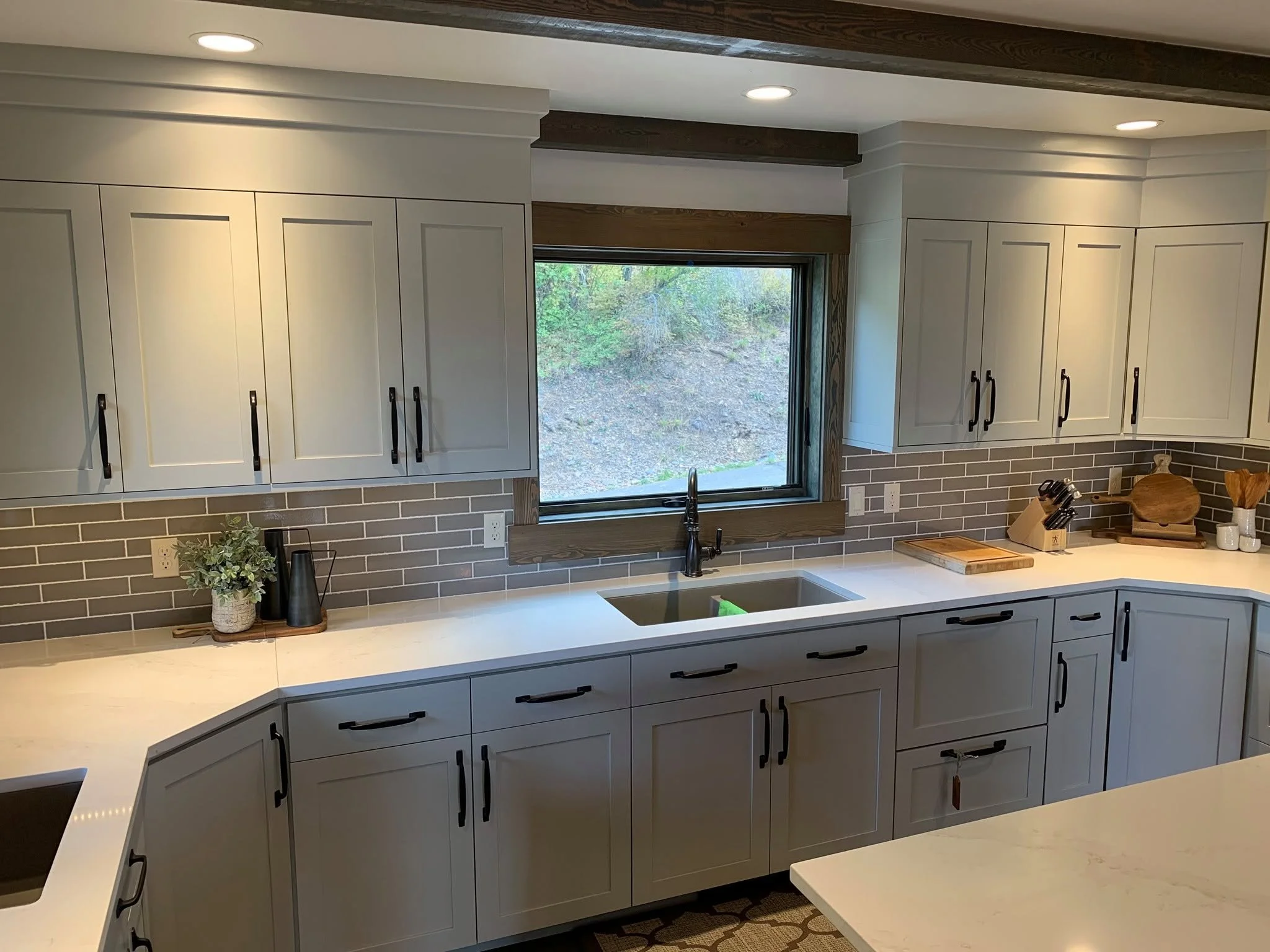 A modern kitchen with white cabinets, black handles, and a gray subway tile backsplash. There is a window above the sink with a view of trees outside. On the countertop are a leafy plant, two black vases, and a knife block with knives. Recessed light