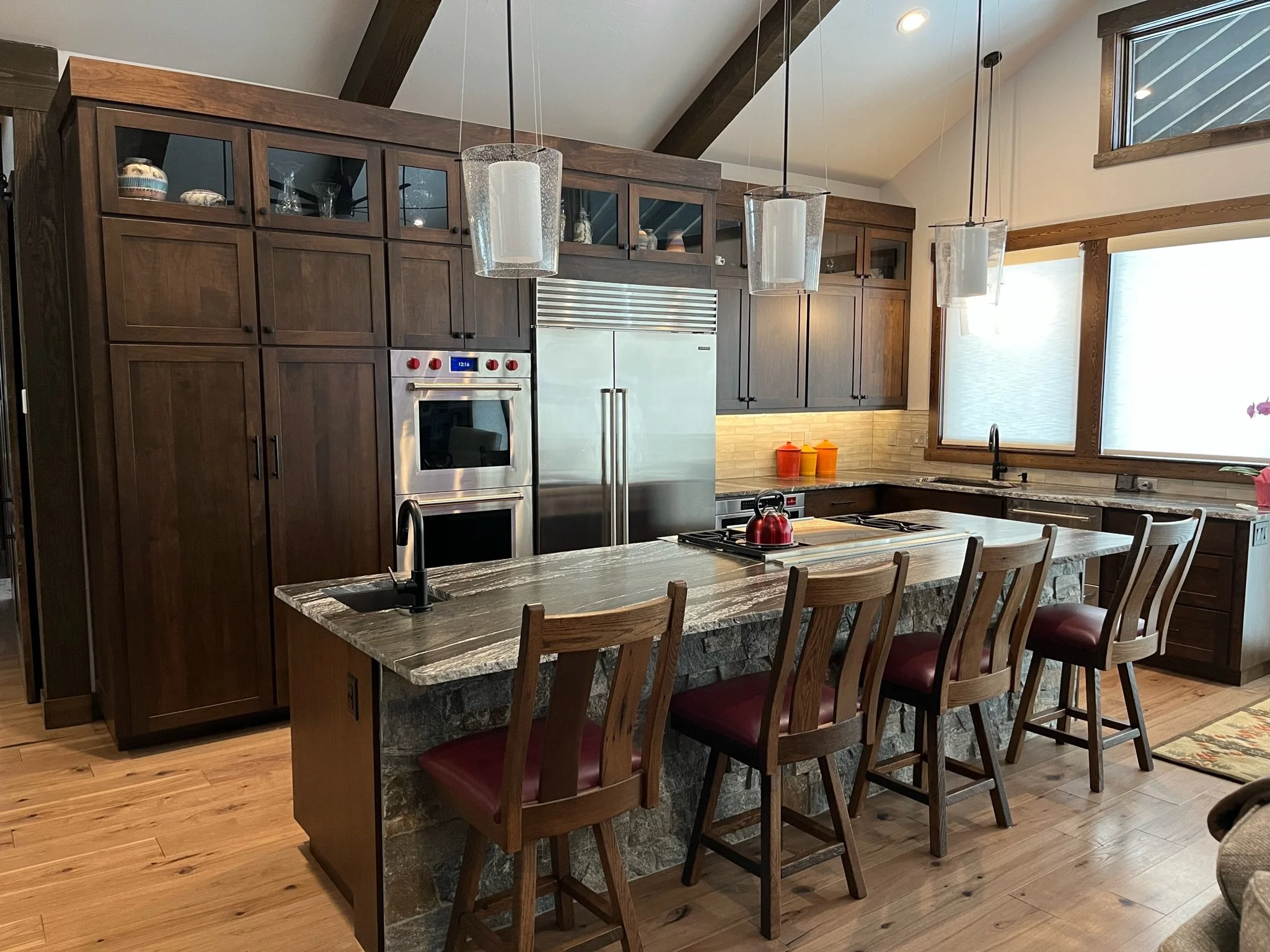 Modern kitchen with dark wood cabinets, granite countertop, stainless steel appliances including a refrigerator and double oven, and hanging pendant lights over a breakfast bar with wooden chairs. Large windows let in natural light.