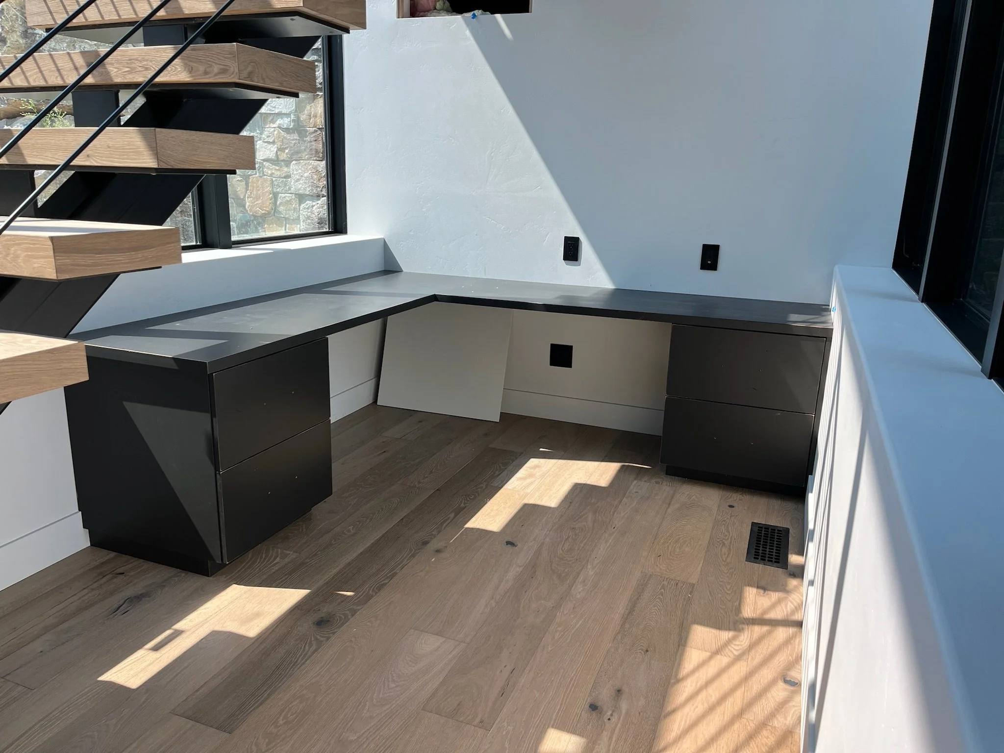 Interior of a modern room with wooden stairs, a corner desk with black and white cabinetry, a large window, and electrical outlets on the wall. Floor is made of wooden planks.