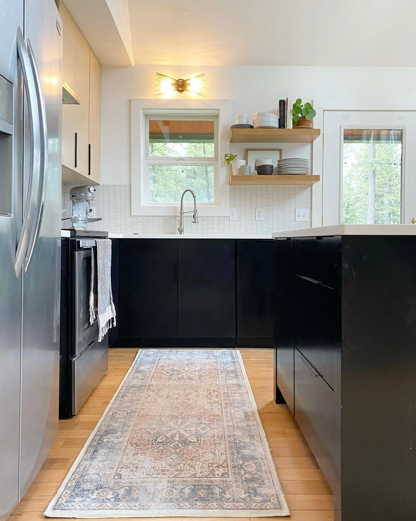 A modern kitchen with black lower cabinets, light upper cabinets, hardwood flooring, a patterned runner rug, a window above the sink, open wooden shelves with dishes and decor, stainless steel appliances, and a glass door leading outside.