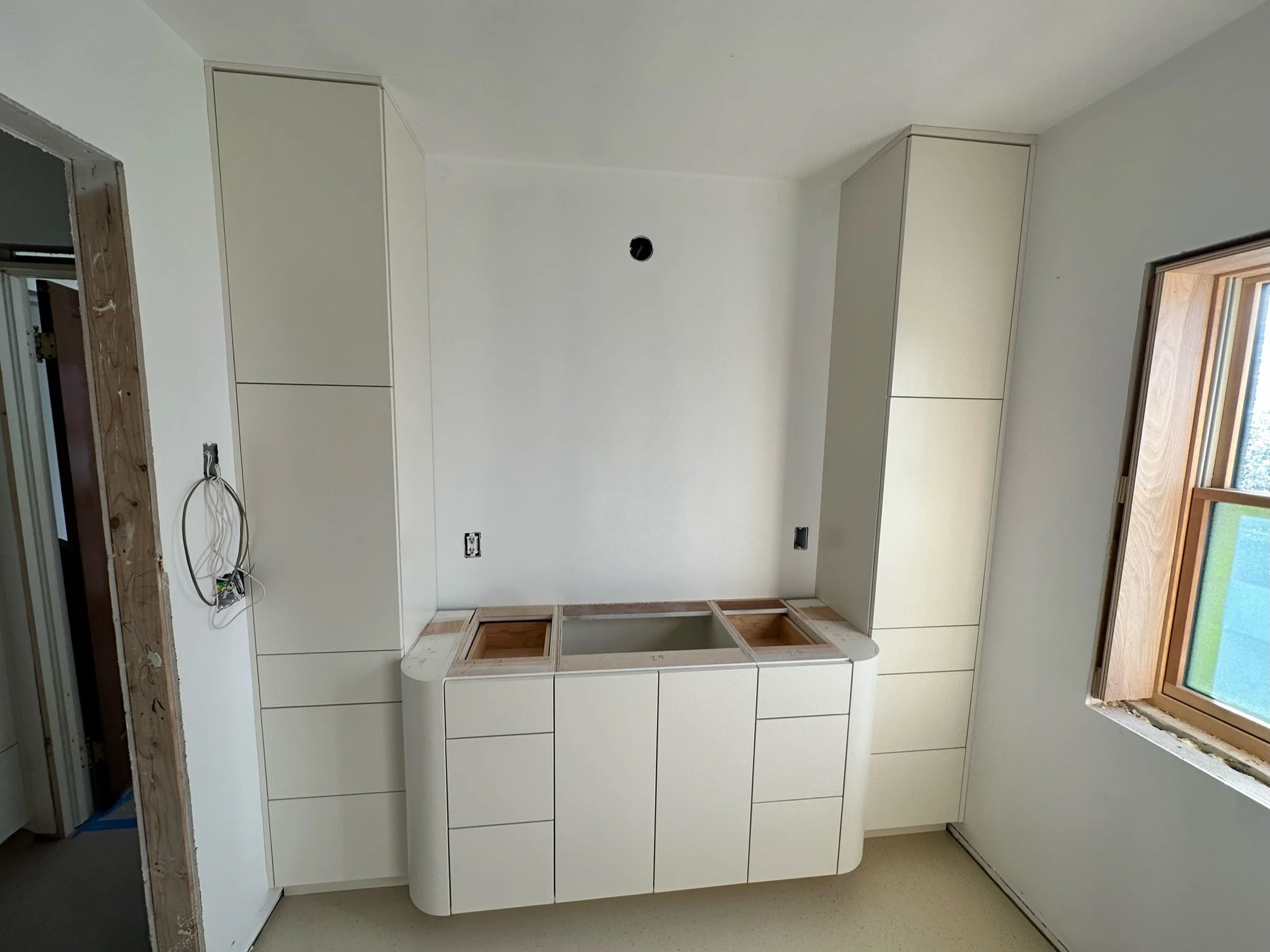 Kitchen under construction with white cabinets and a window, unfinished wall with exposed wiring, and a white countertop with an empty space for a sink.