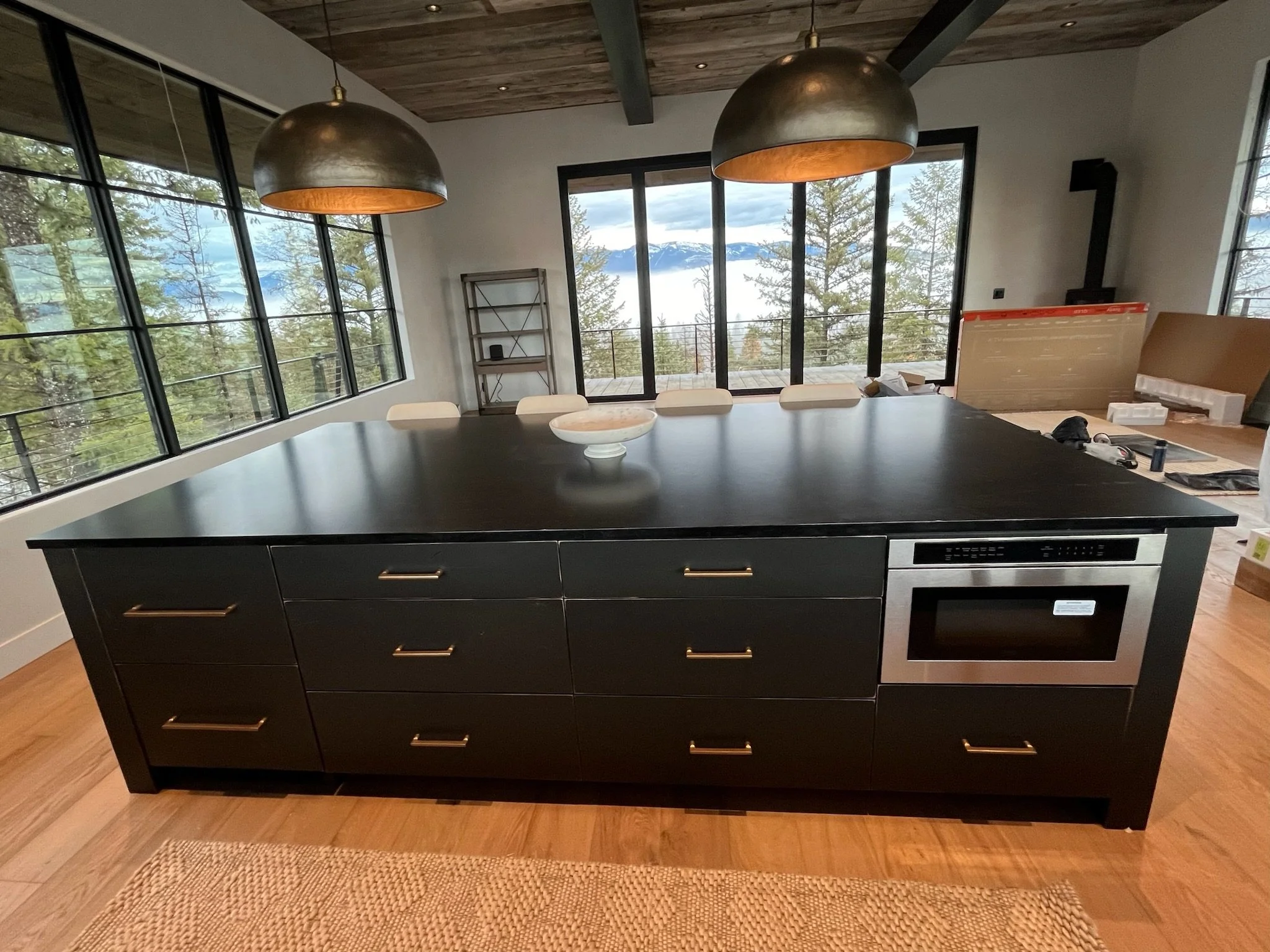 A modern kitchen with black cabinets, a large black island. Hanging pendant lights above the island, large windows providing a mountain view, and a sliding glass door leading to a balcony with trees and mountains in the distance.